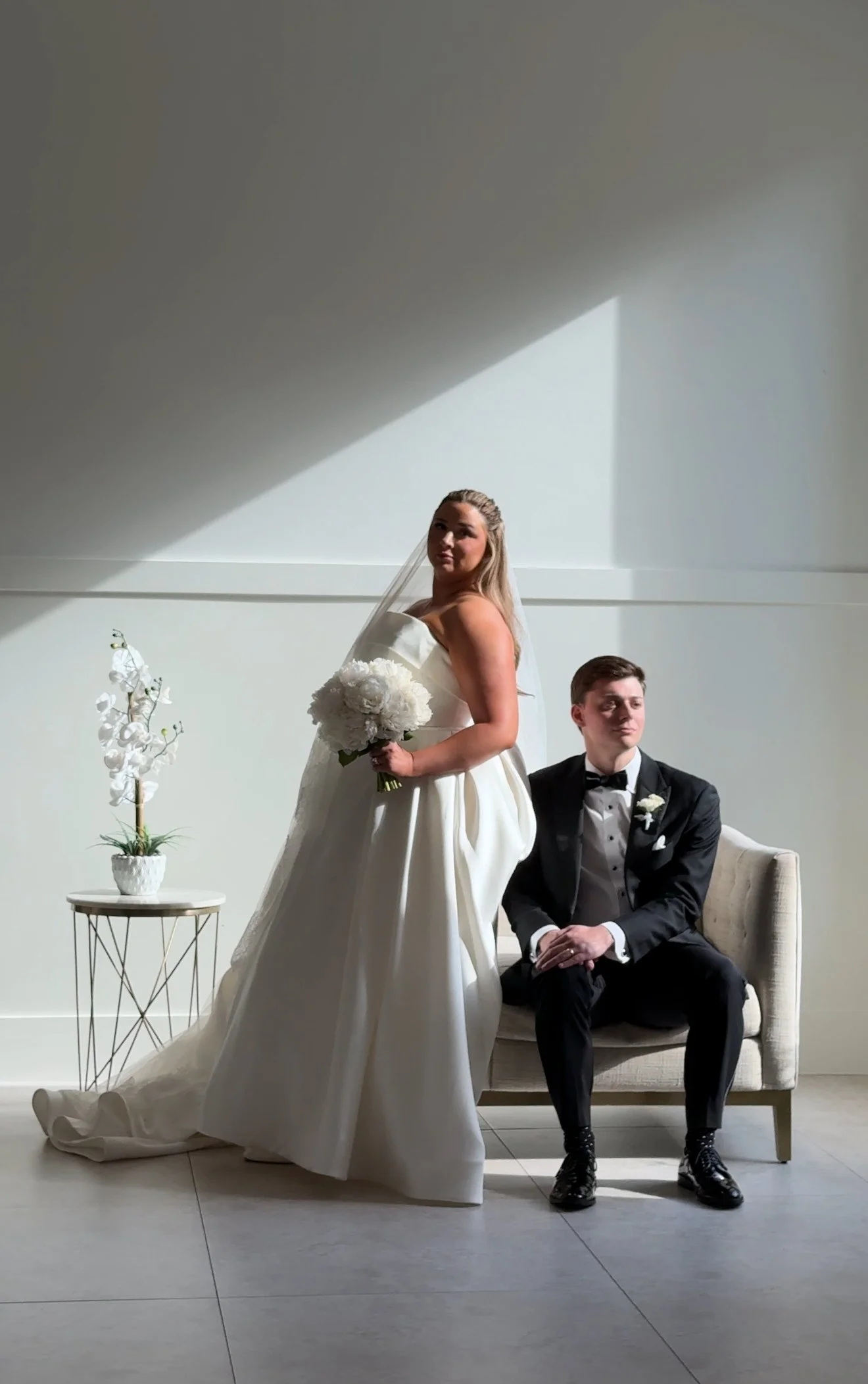 A bride and groom pose in a minimalistic, well-lit room. The bride is standing, holding a bouquet of white flowers, and wearing a strapless white wedding gown. The groom is sitting on a light-colored sofa, dressed in a tuxedo with a bow tie, looking to the side. A small table with a plant is nearby, against a plain wall with a shadow cast on it.