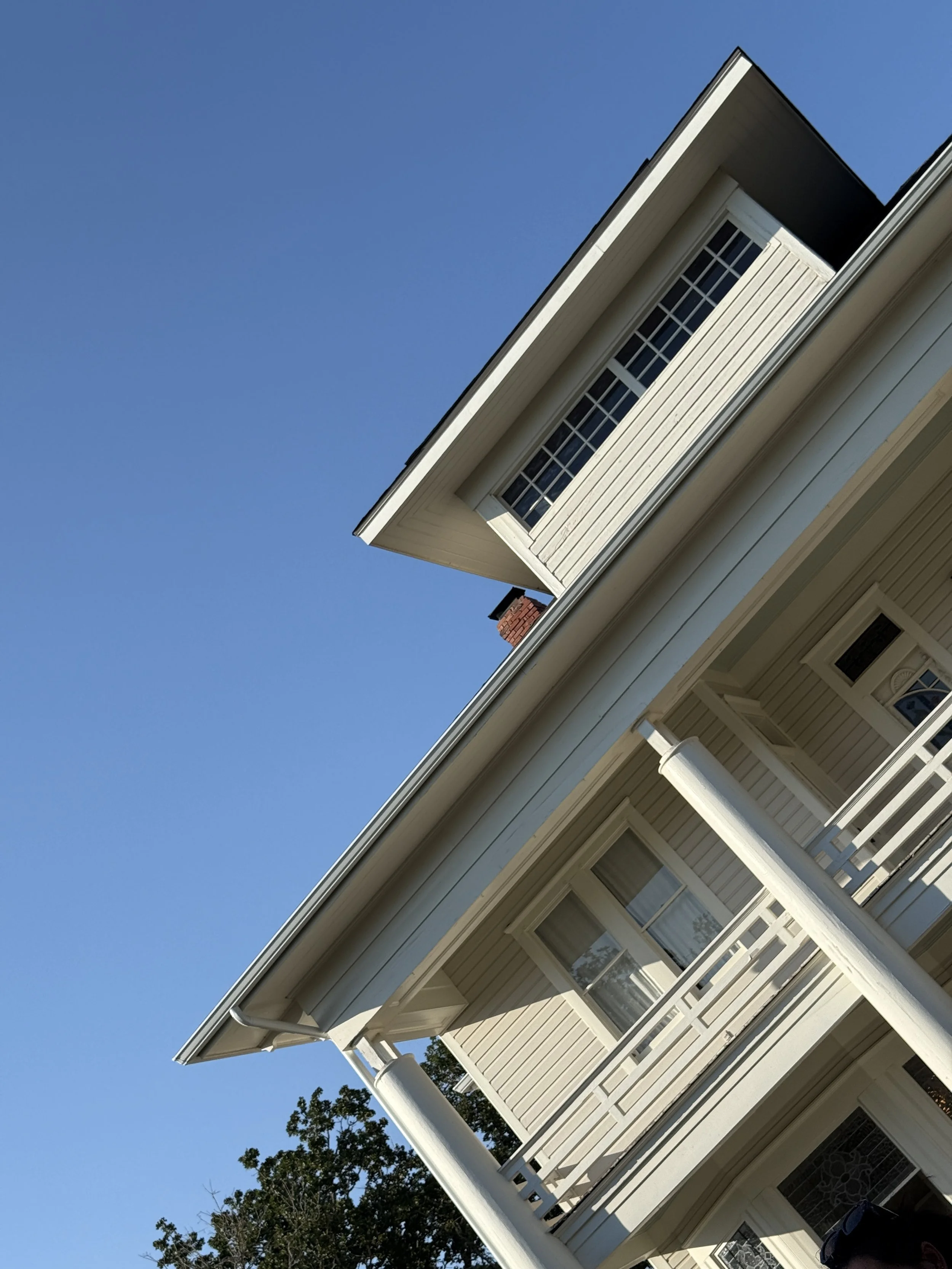 Side view of a light-colored house with white porch railings and windows, against a clear blue sky.