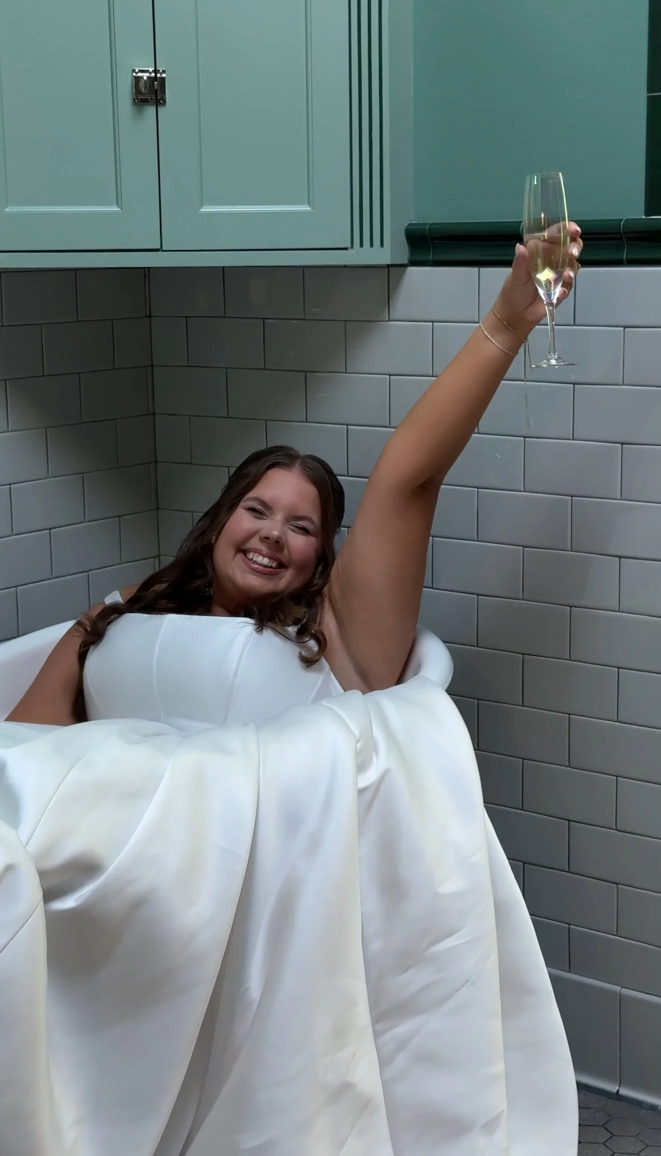 A woman in a white dress, smiling and reclining in a bathtub, holding a glass of champagne raised in celebration.