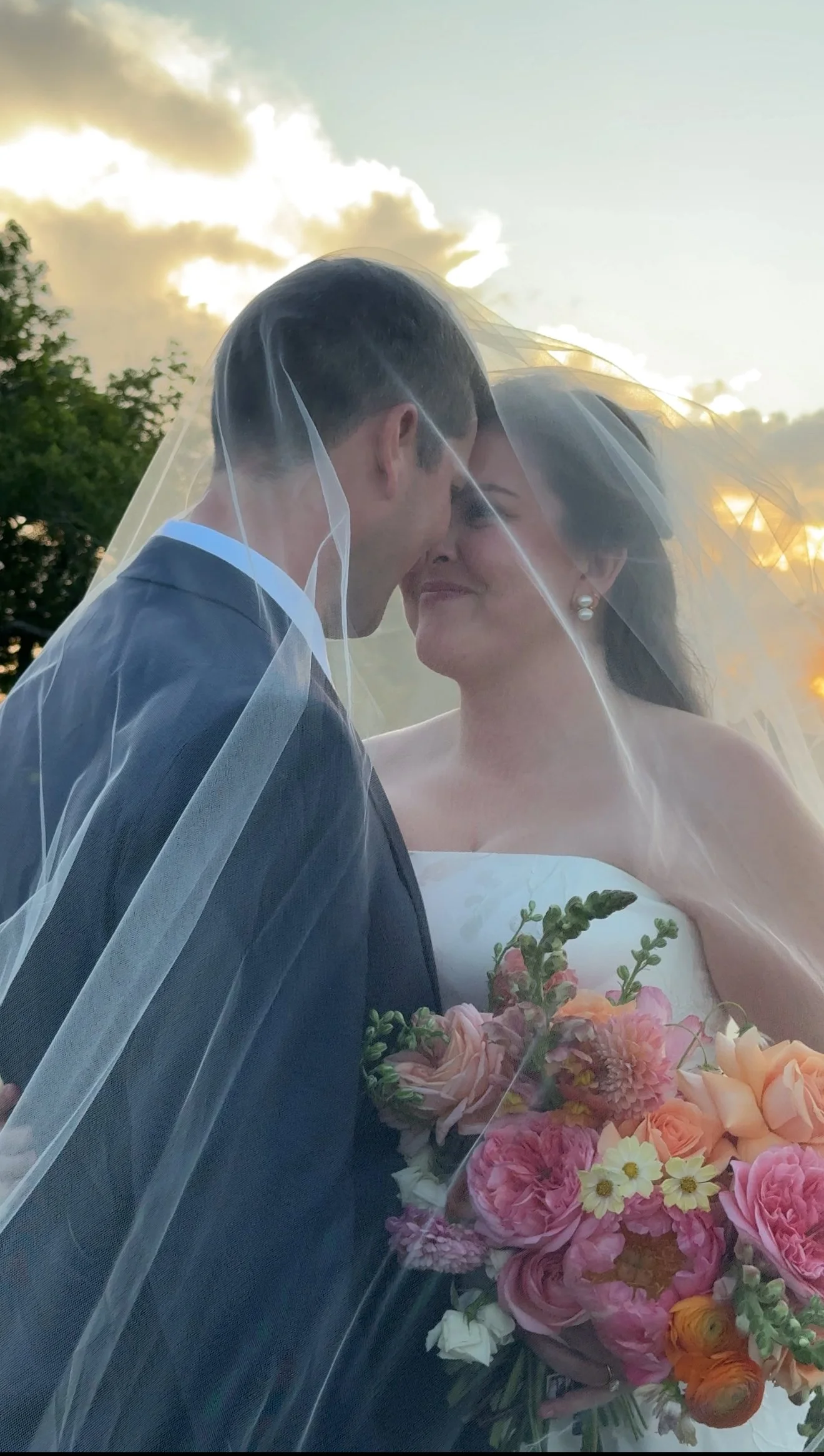 A newlywed couple stands close under a sheer veil, smiling lovingly at each other during sunset. The bride holds a colorful bouquet of flowers.