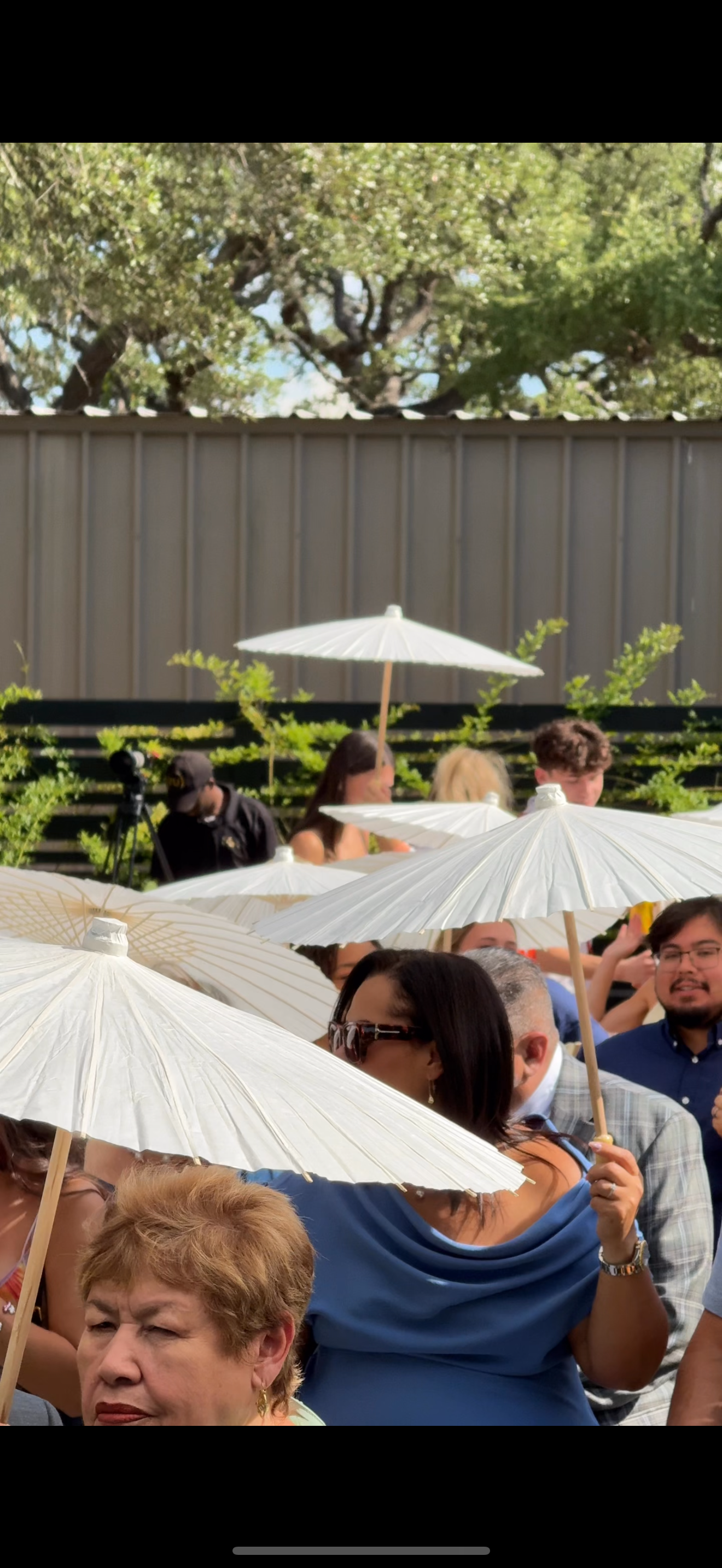 People seated outdoors at a sunny event, holding white parasols for shade, with trees and a metal fence in the background.