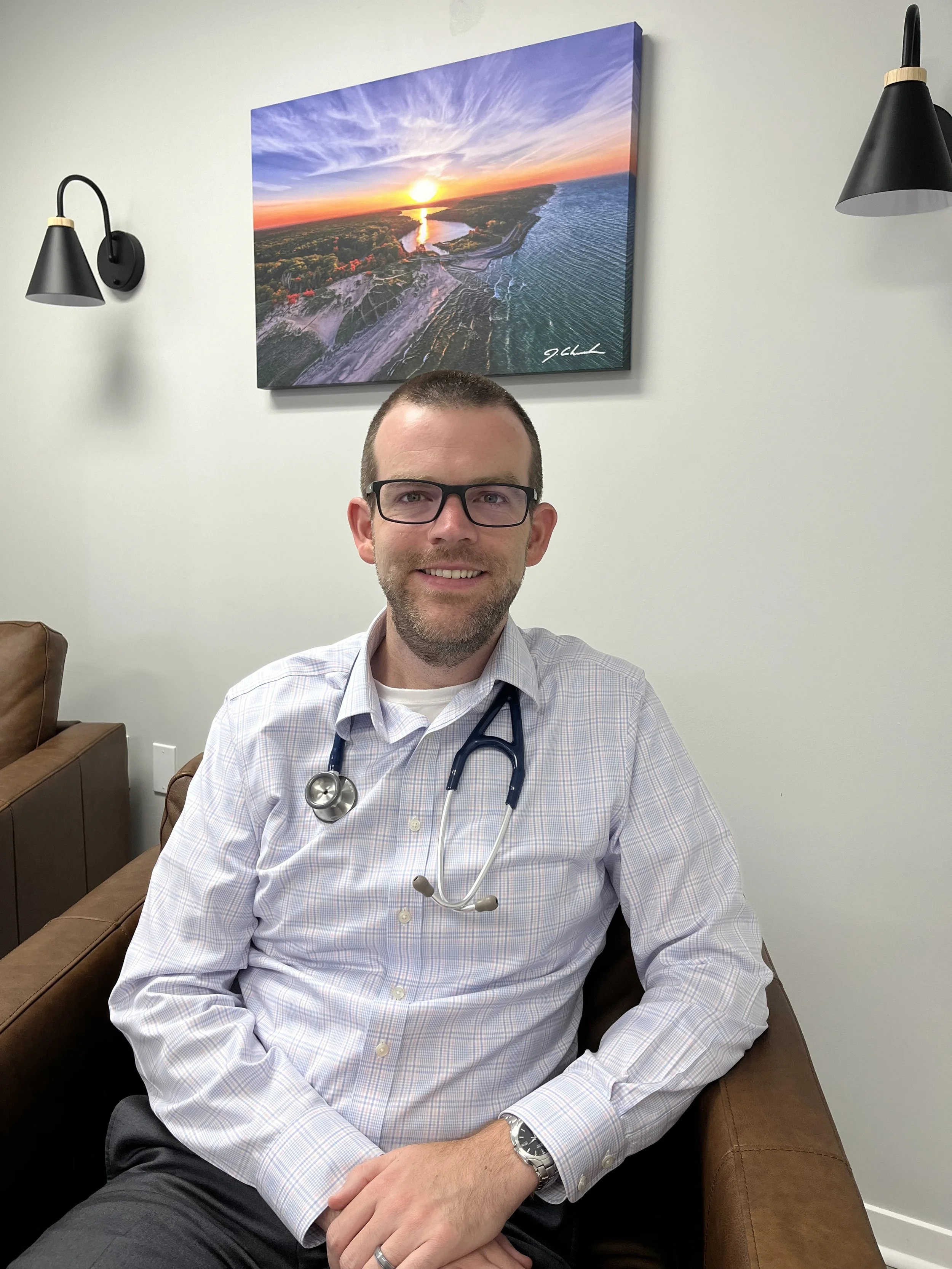 Greg Holton male doctor with glasses, wearing a stethoscope around his neck, sitting on a brown leather chair in an office. Behind him is a large landscape photograph of a sunset over a river and coastline, on a white wall with black wall sconces.