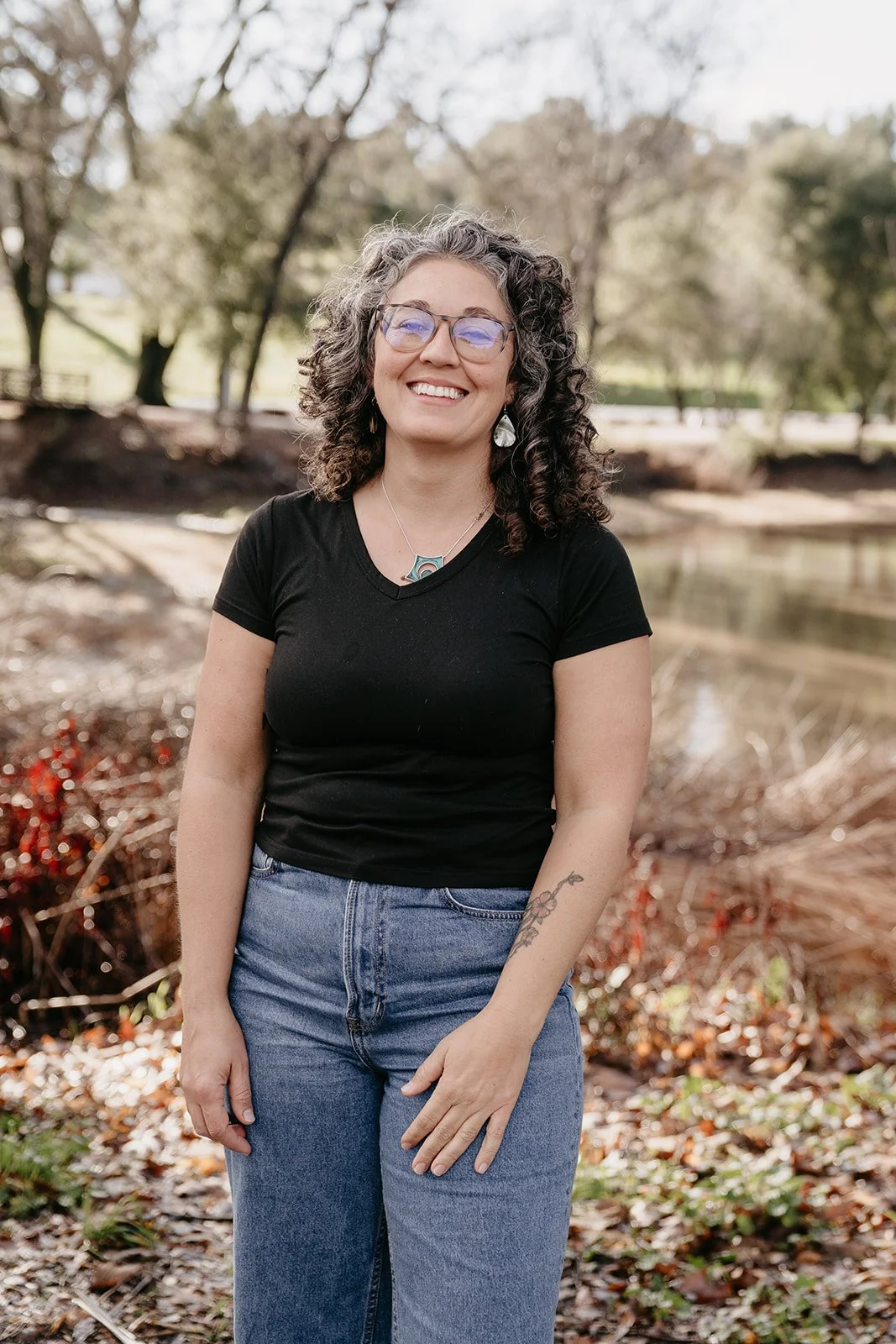 Smiling woman with curly hair, glasses, black t-shirt, and jeans standing outdoors near a pond with trees.
