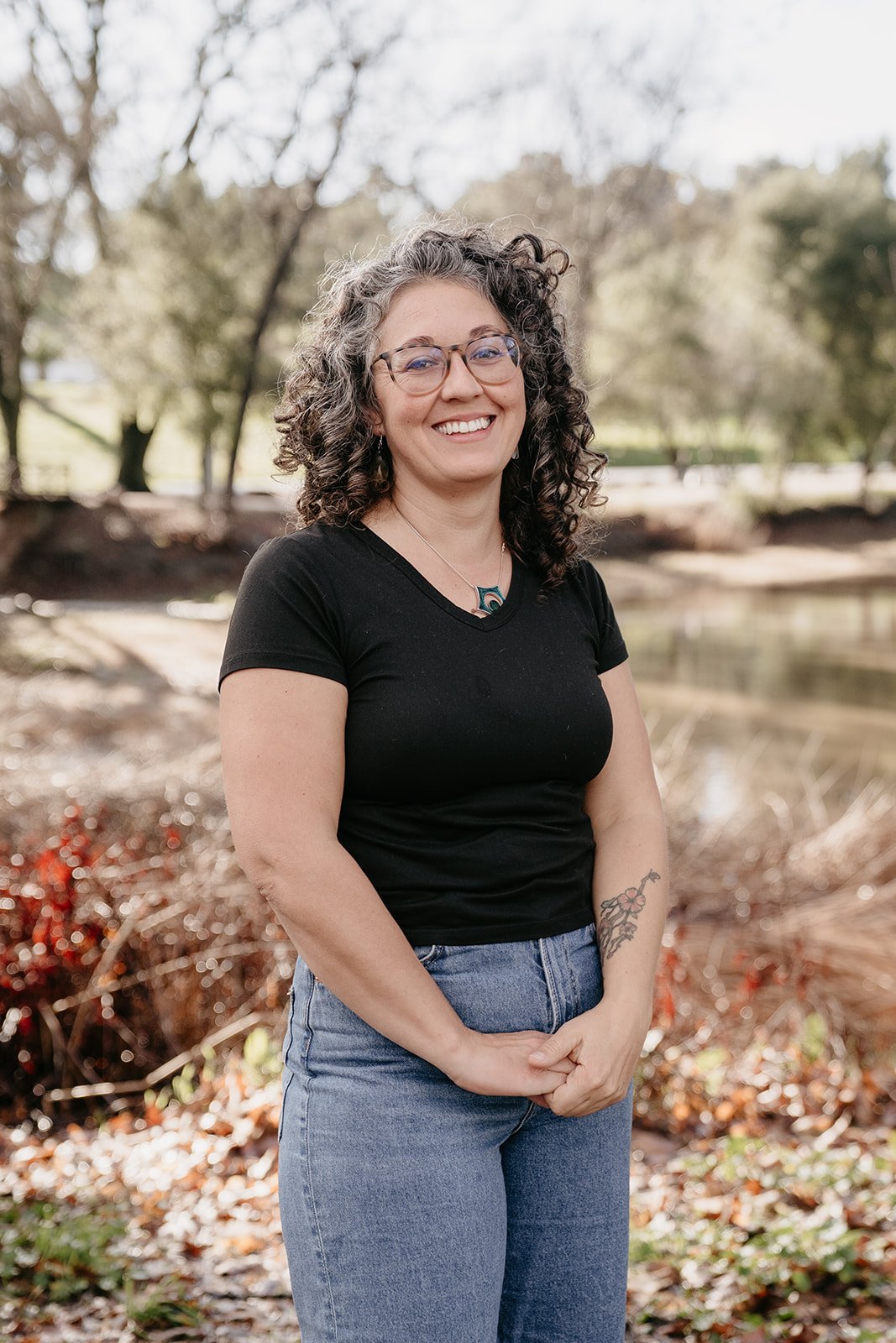 A woman with curly hair and glasses smiling outdoors near a pond, wearing a black t-shirt and blue jeans.