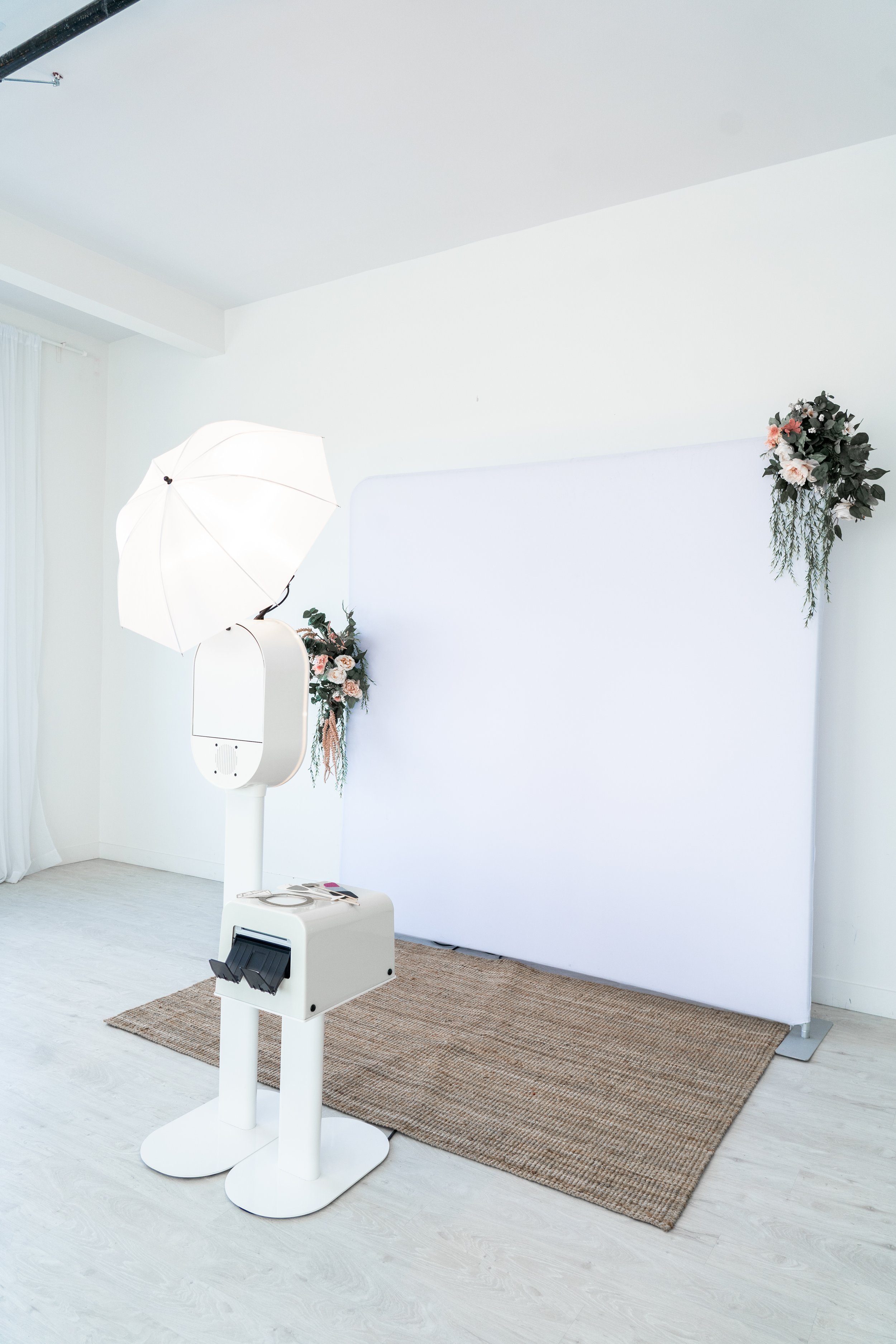 Photo of a photobooth setup with a white backdrop decorated with pink and white flowers, a white umbrella, and a photo printer in a bright, minimalistic room.
