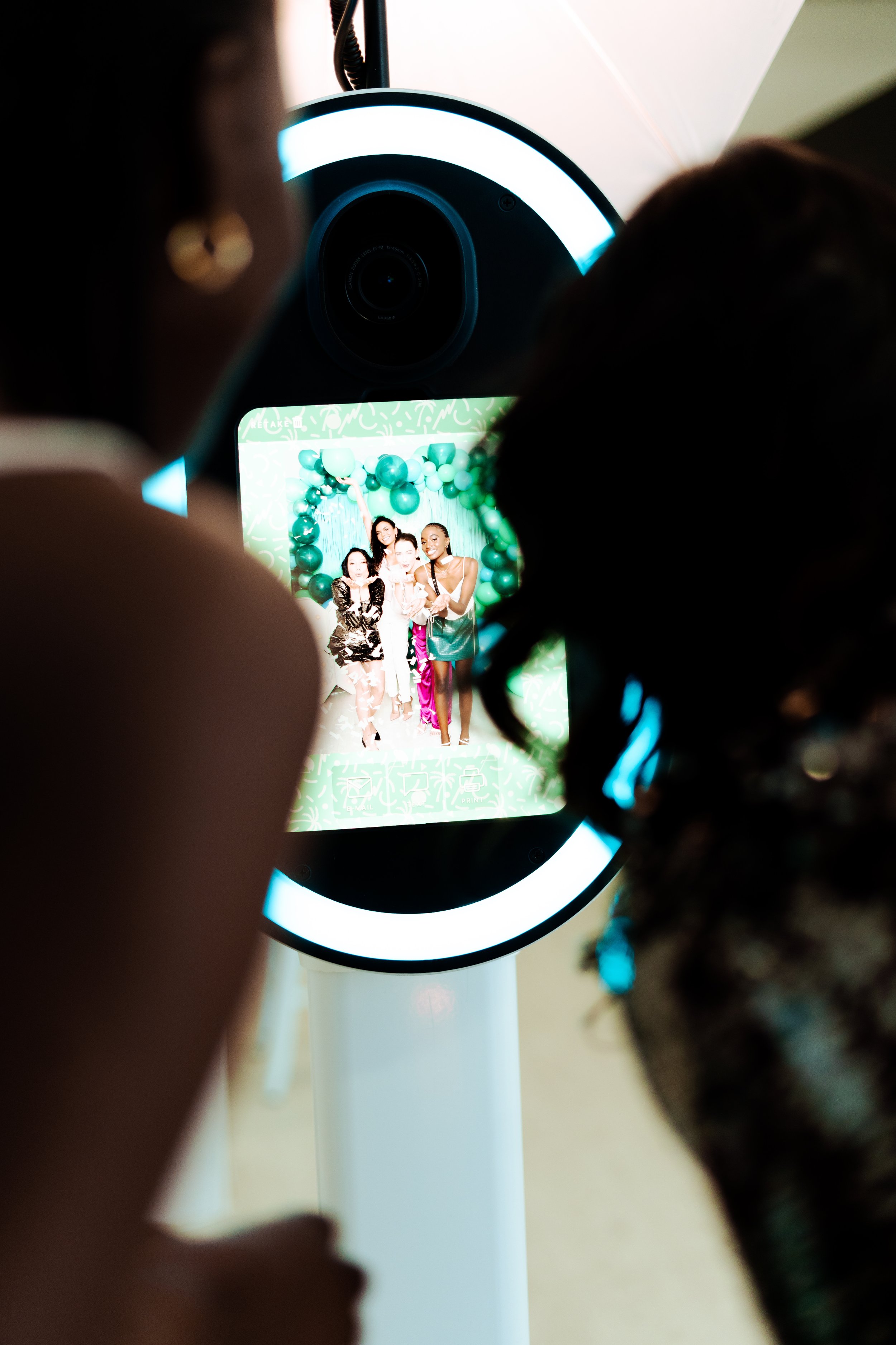 Two women and three children taking a group photo with a photobooth mirror decorated with green balloons.