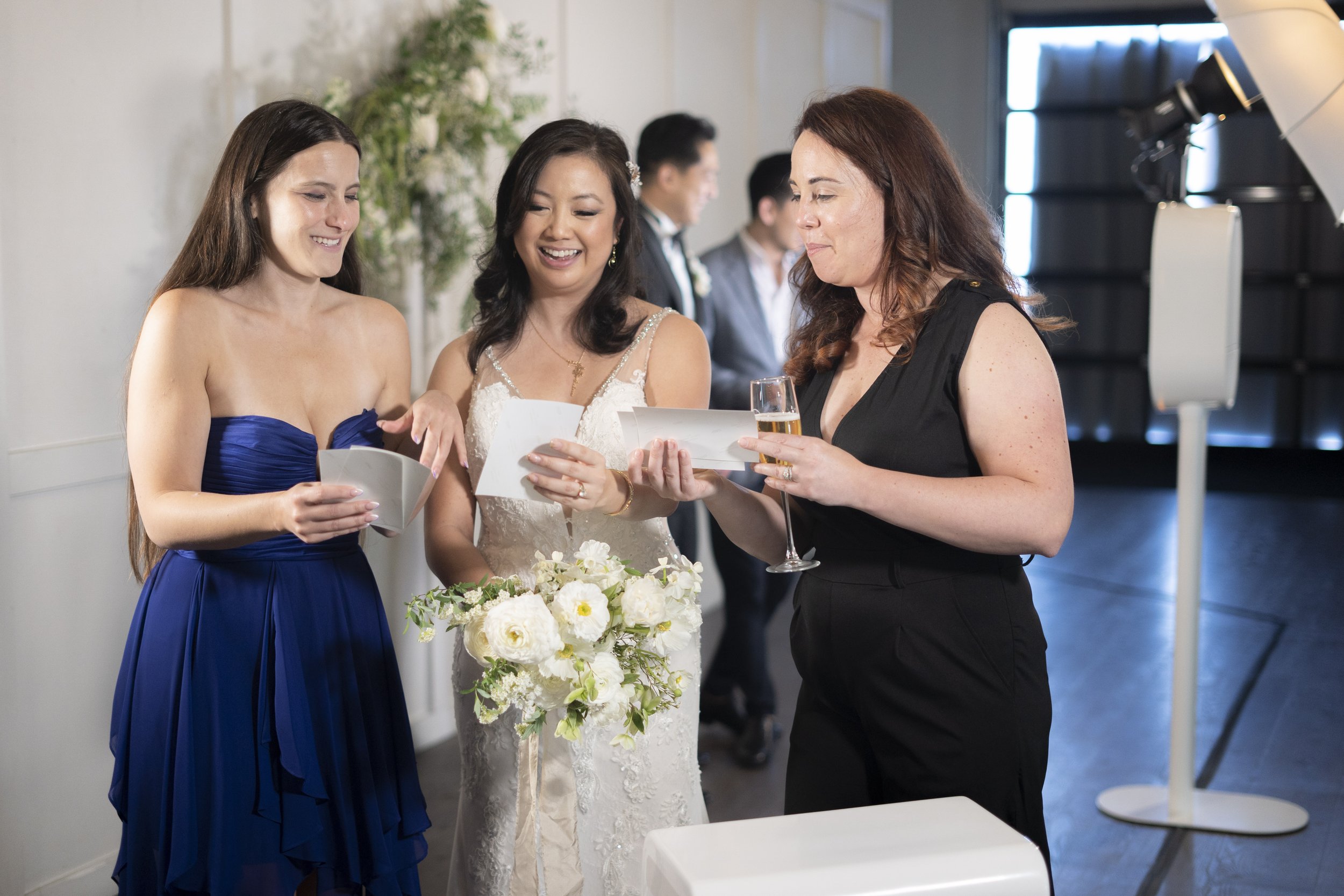 Three women at a wedding reception, reading cards or notes, with one woman holding a glass of champagne, smiling and interacting with each other.