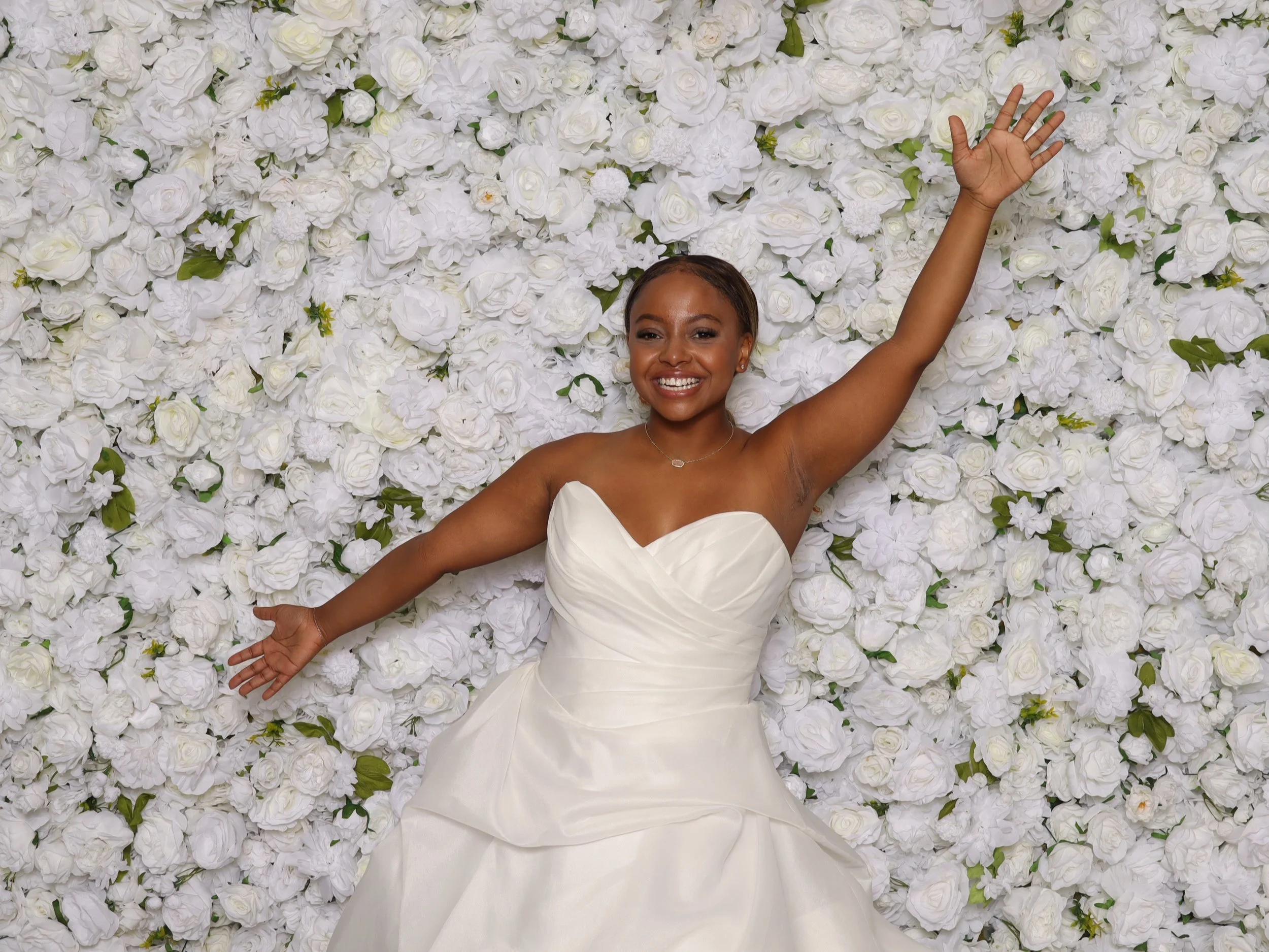 A woman in a white wedding dress smiling and lying on a bed of white roses with her arms raised.