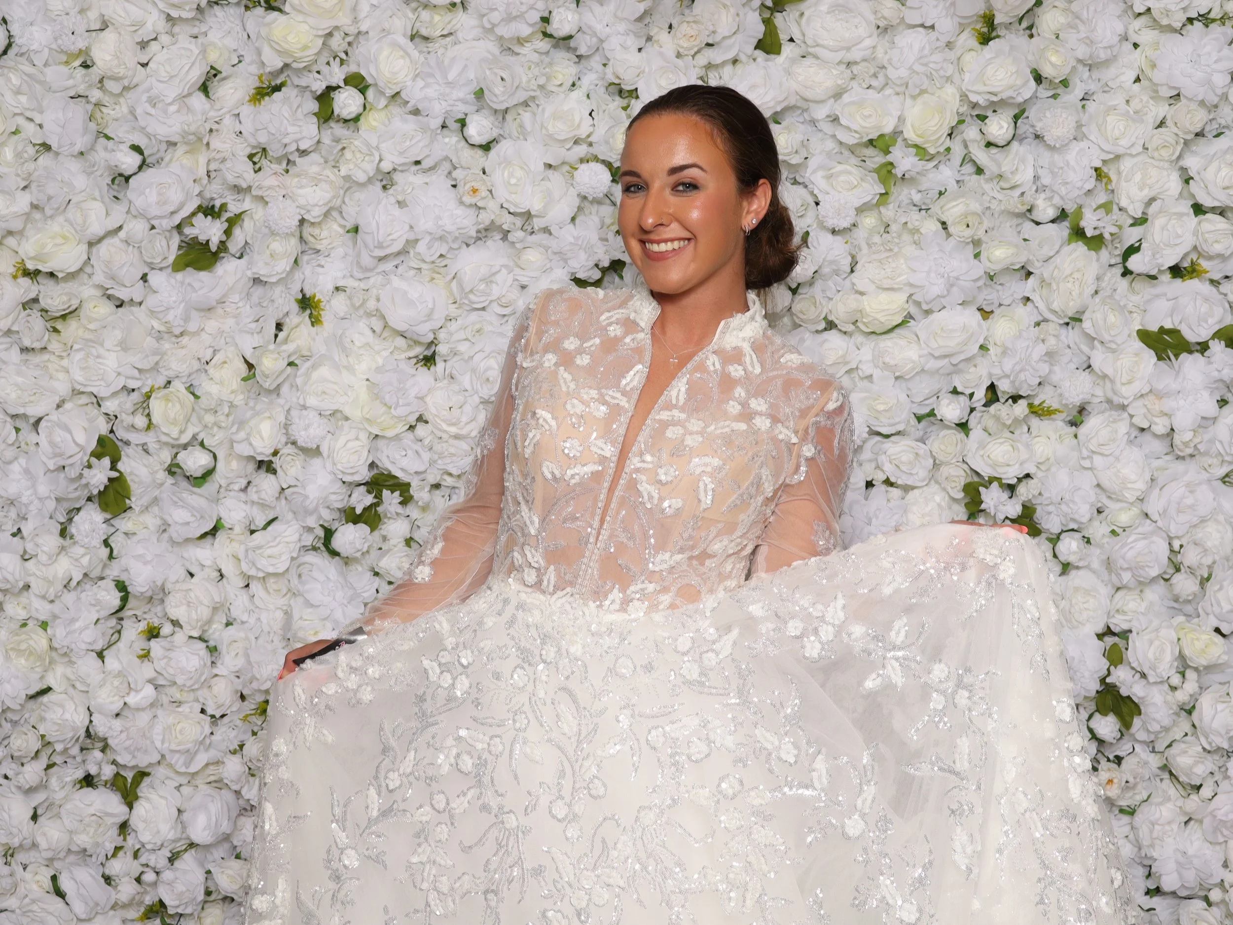 A young woman in a wedding dress stands in front of a wall of white flowers, smiling, holding her dress up slightly. She has brown hair and is wearing earrings.