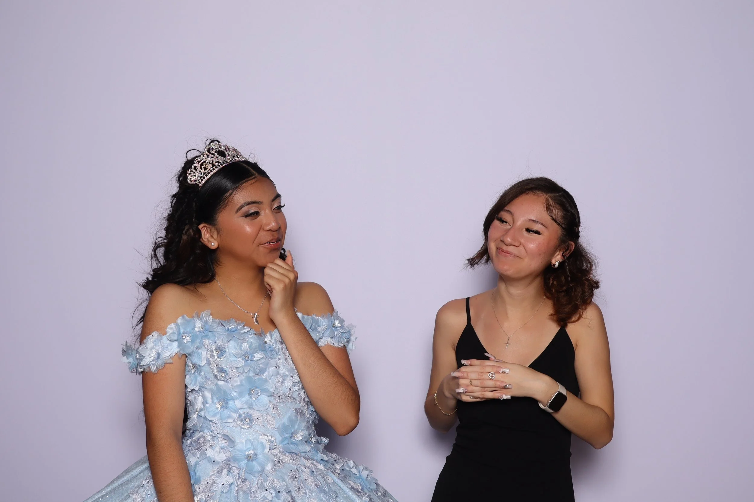 A young woman wearing a light blue floral dress and a tiara, possibly a prom queen or bride, standing next to another woman in a black dress, both smiling and engaging in conversation against a plain background.
