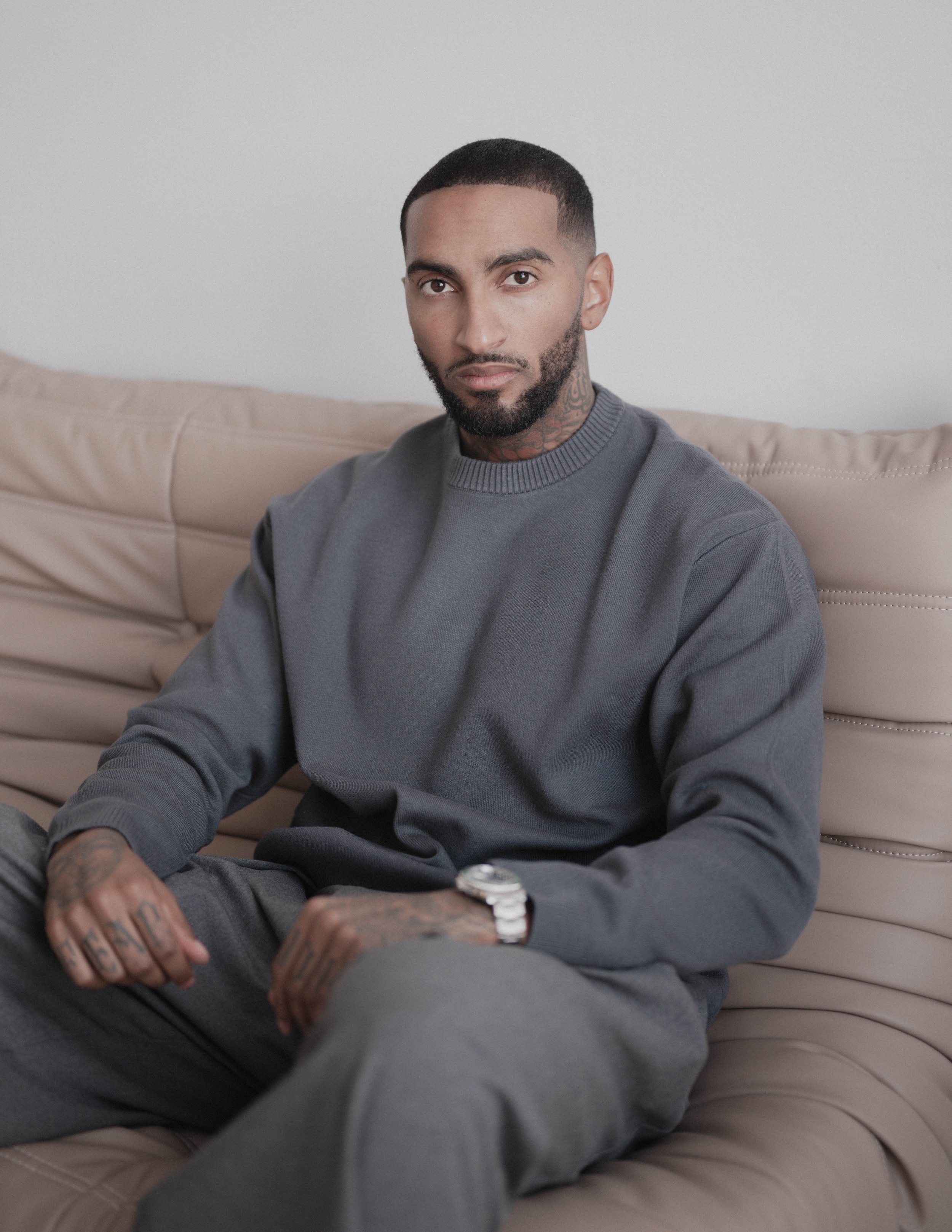 Un homme assis sur un canapé beige, portant un sweat gris foncé, un pantalon gris, une montre en métal, avec des tatouages visibles sur les mains et le cou, regardant vers l'objectif.