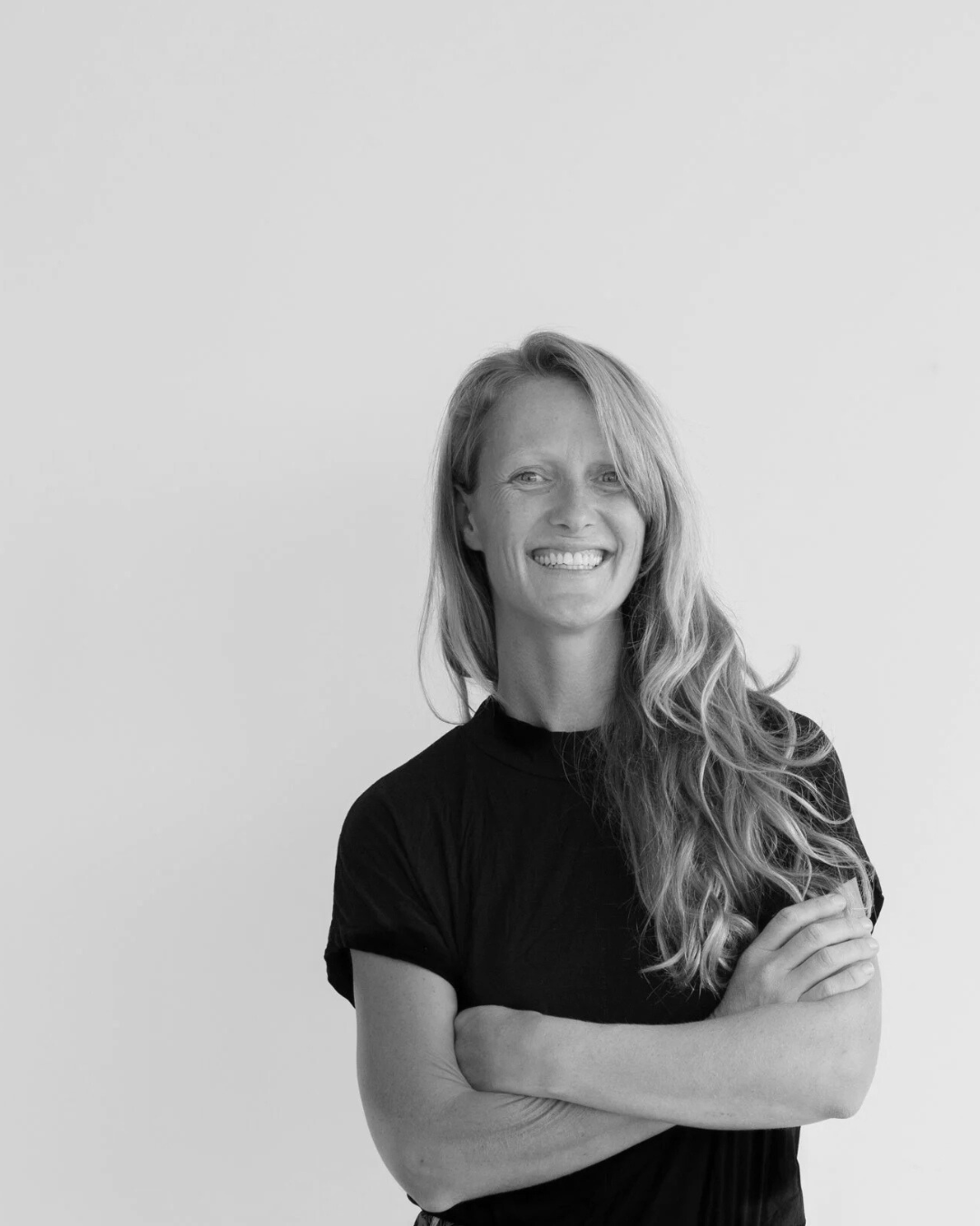 A woman with long, wavy blonde hair wearing a black shirt, smiling against a plain background.