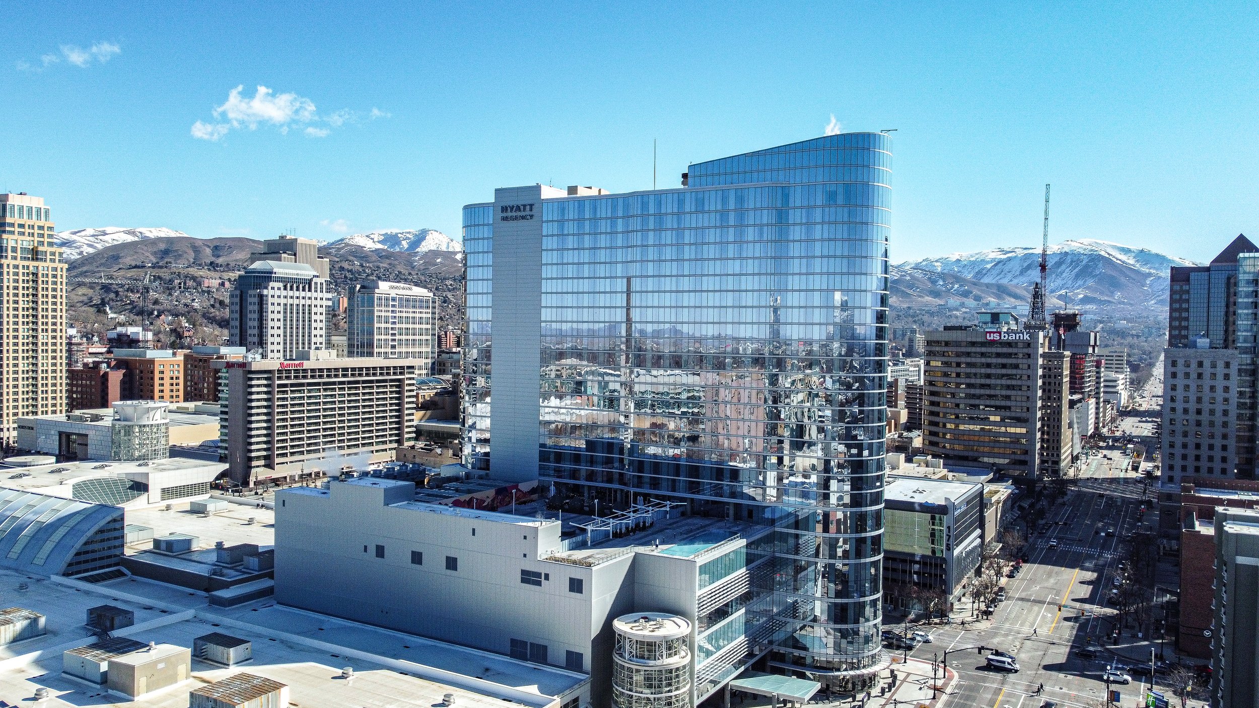 Front view of a two-story white house with large arched windows, a garage, and a landscaped yard with bushes, grass, and stairs, with neighboring houses and mountains in the background.