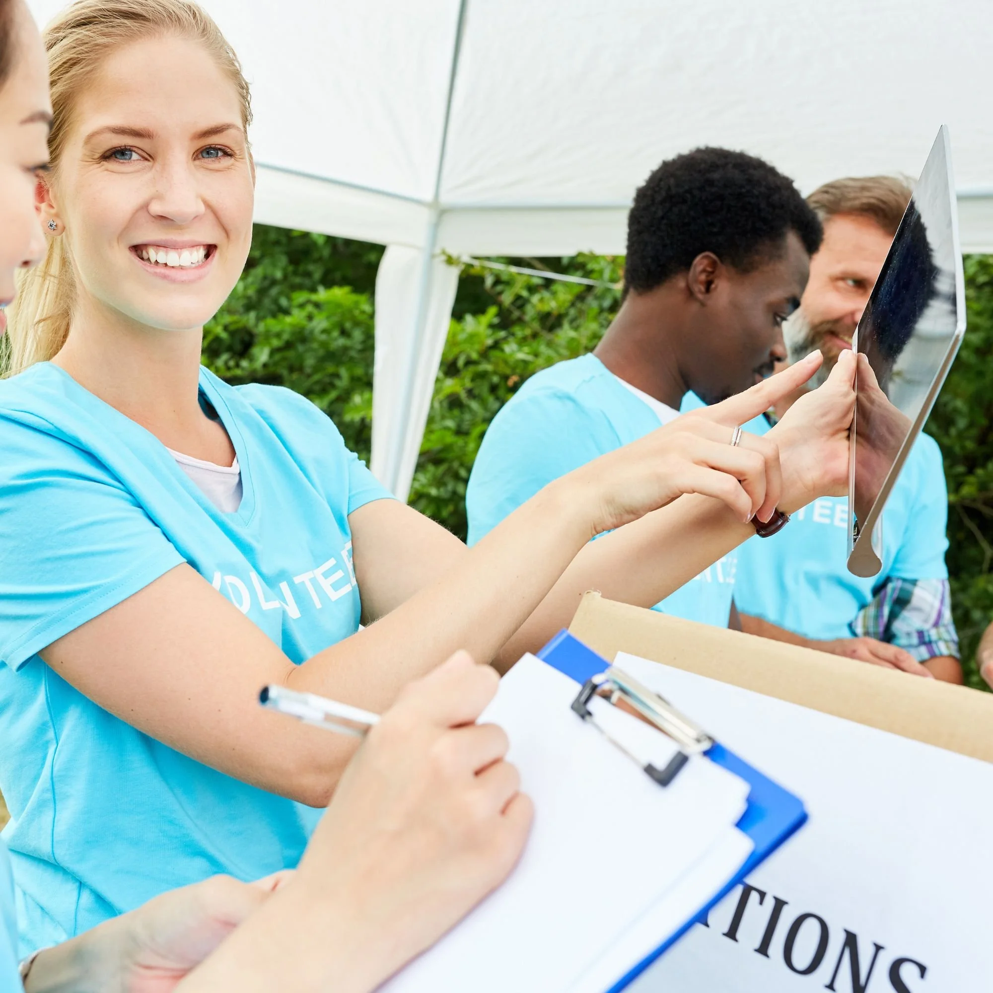 Smiling woman with blonde hair in blue shirt at voting location, with other people holding a tablet and writing on clipboard.