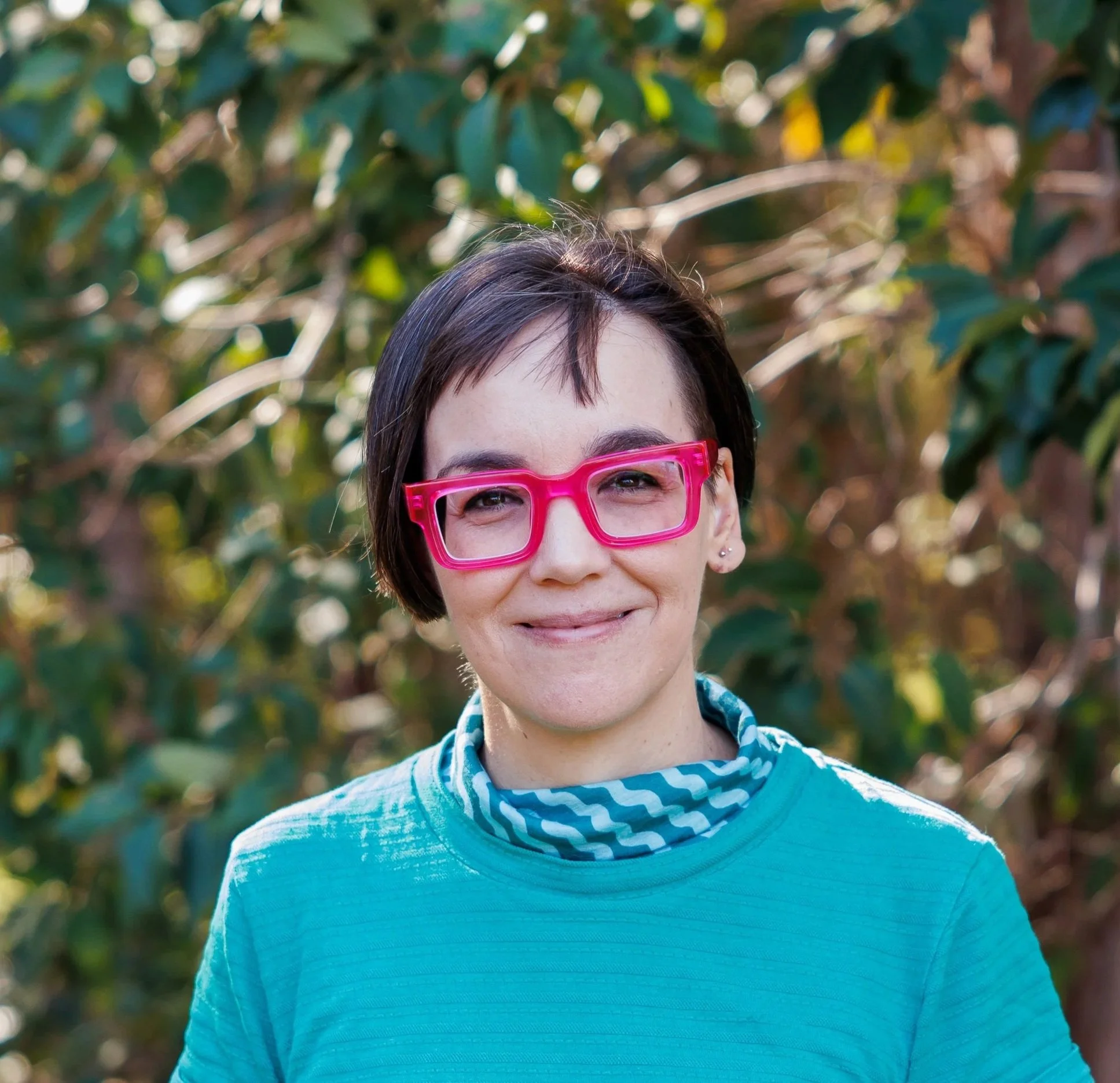 A woman with short dark hair, wearing tortoiseshell glasses and a denim shirt, smiling against a brown background.