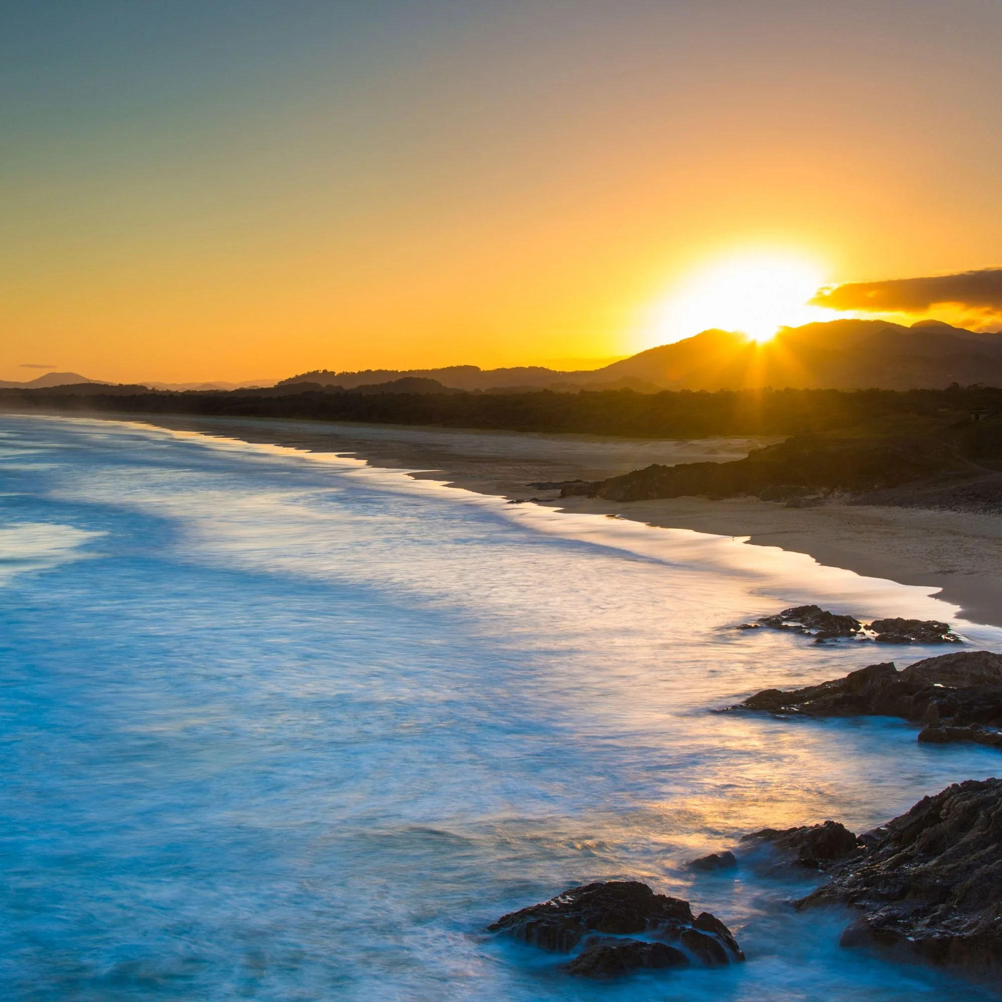 Sunset over the ocean with waves crashing on rocks along the shoreline, with mountains in the distance.