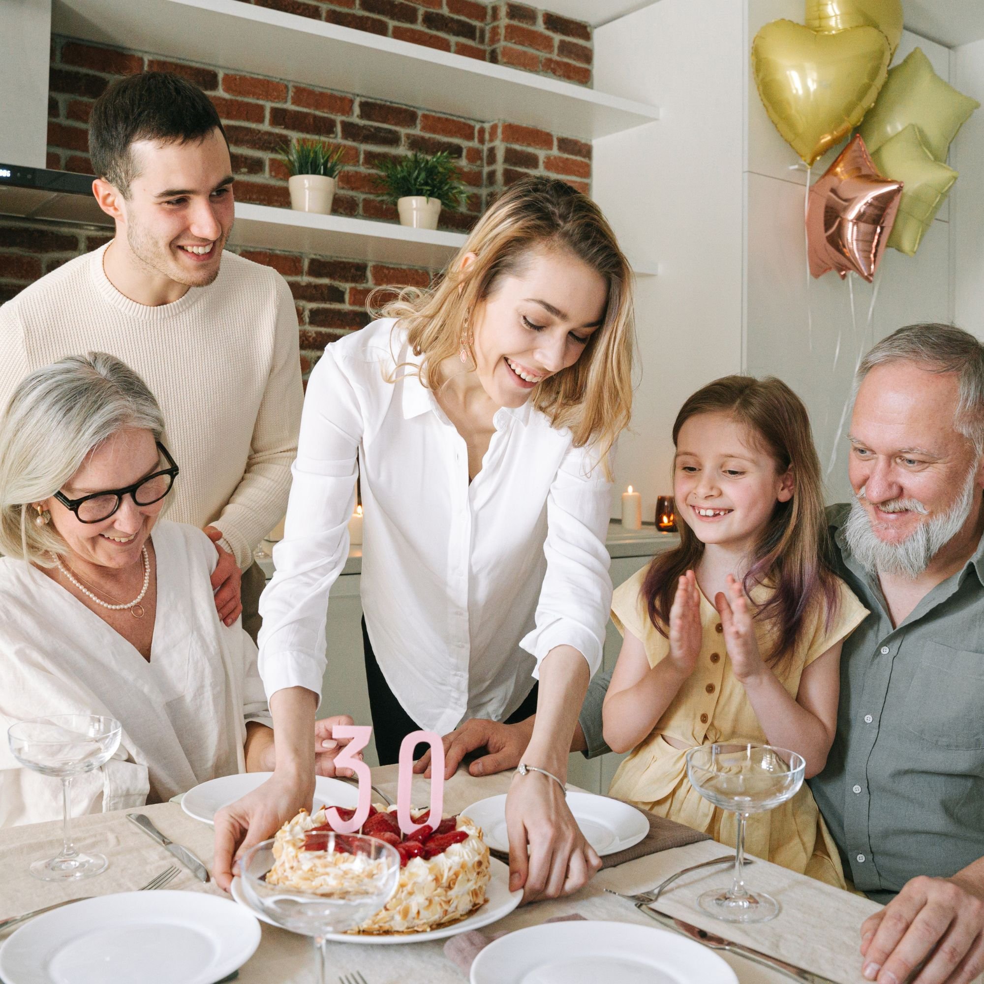 Group of people celebrating a birthday around a table with a cake, candle, and balloons in a cozy indoor setting.