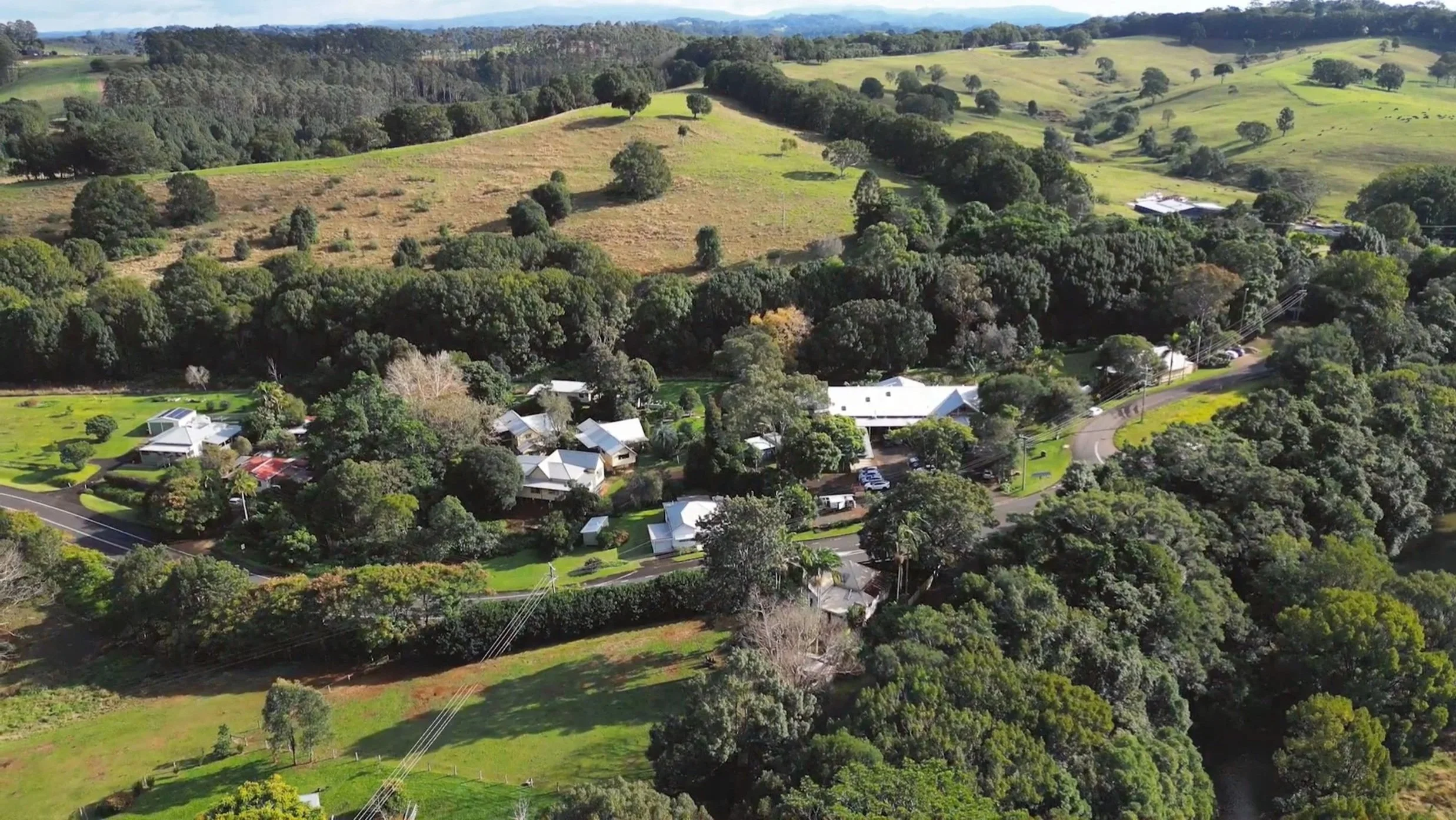 Aerial view of a rural landscape featuring green hills, scattered trees, a residential area with houses, and winding roads.