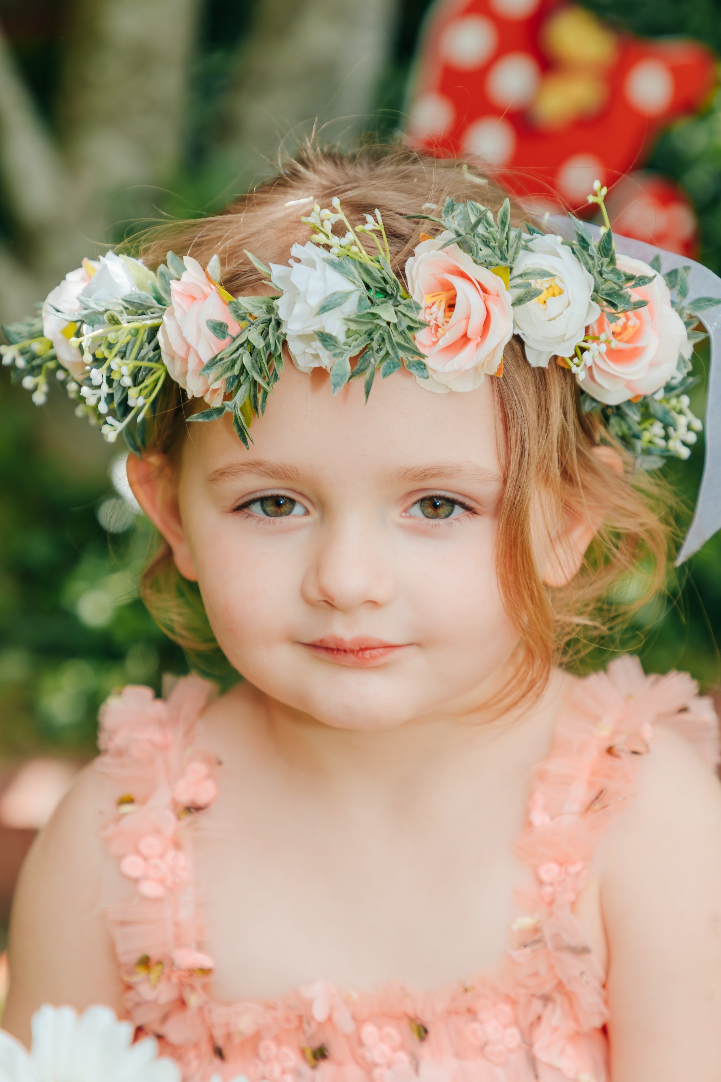 A young girl with light brown hair and green eyes wearing a pink dress with floral embellishments and a flower crown made of roses and greenery. Memphis and Hernando Family Photographer.