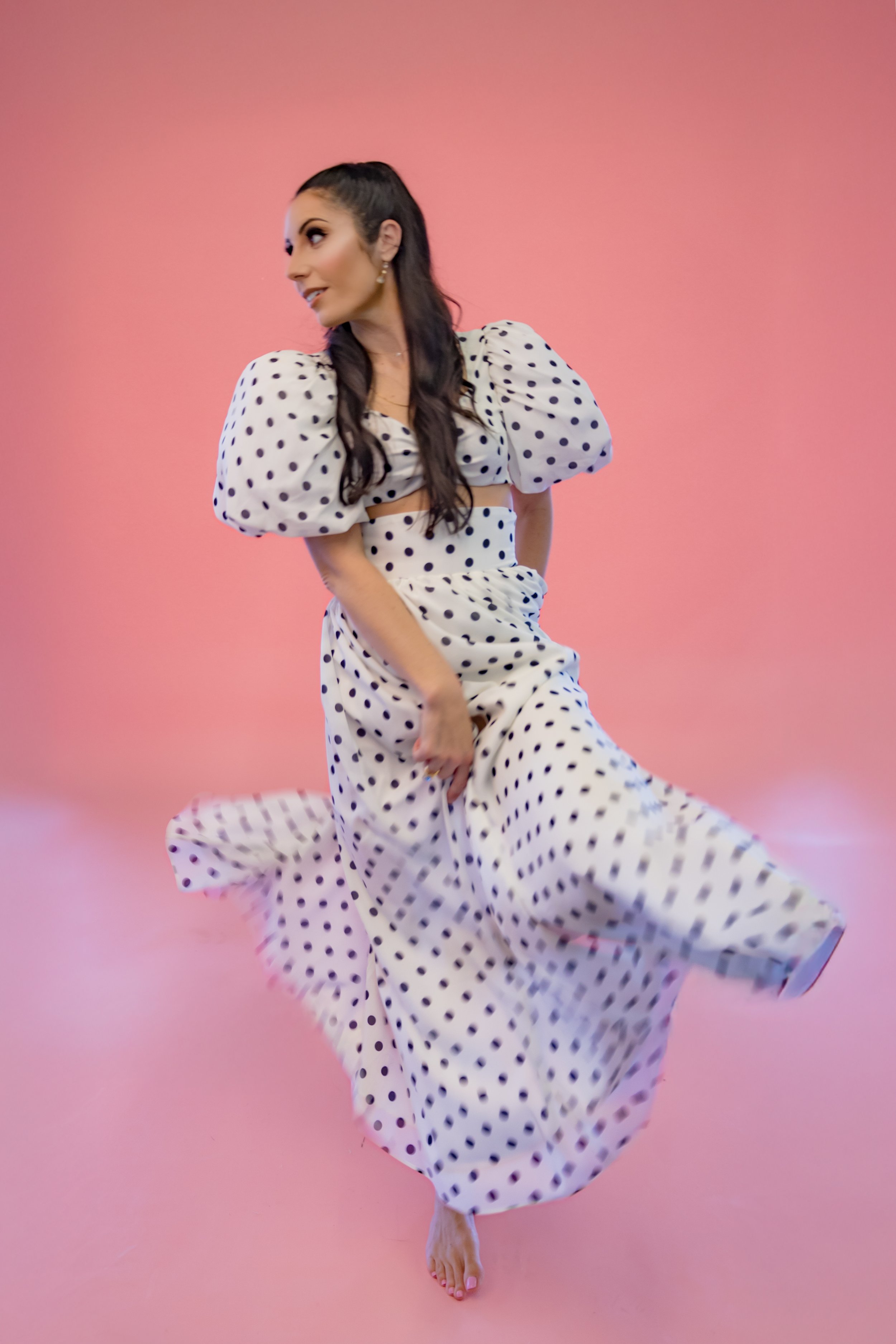 A woman dressed in a white polka dot dress with puffed sleeves standing on a pink background.