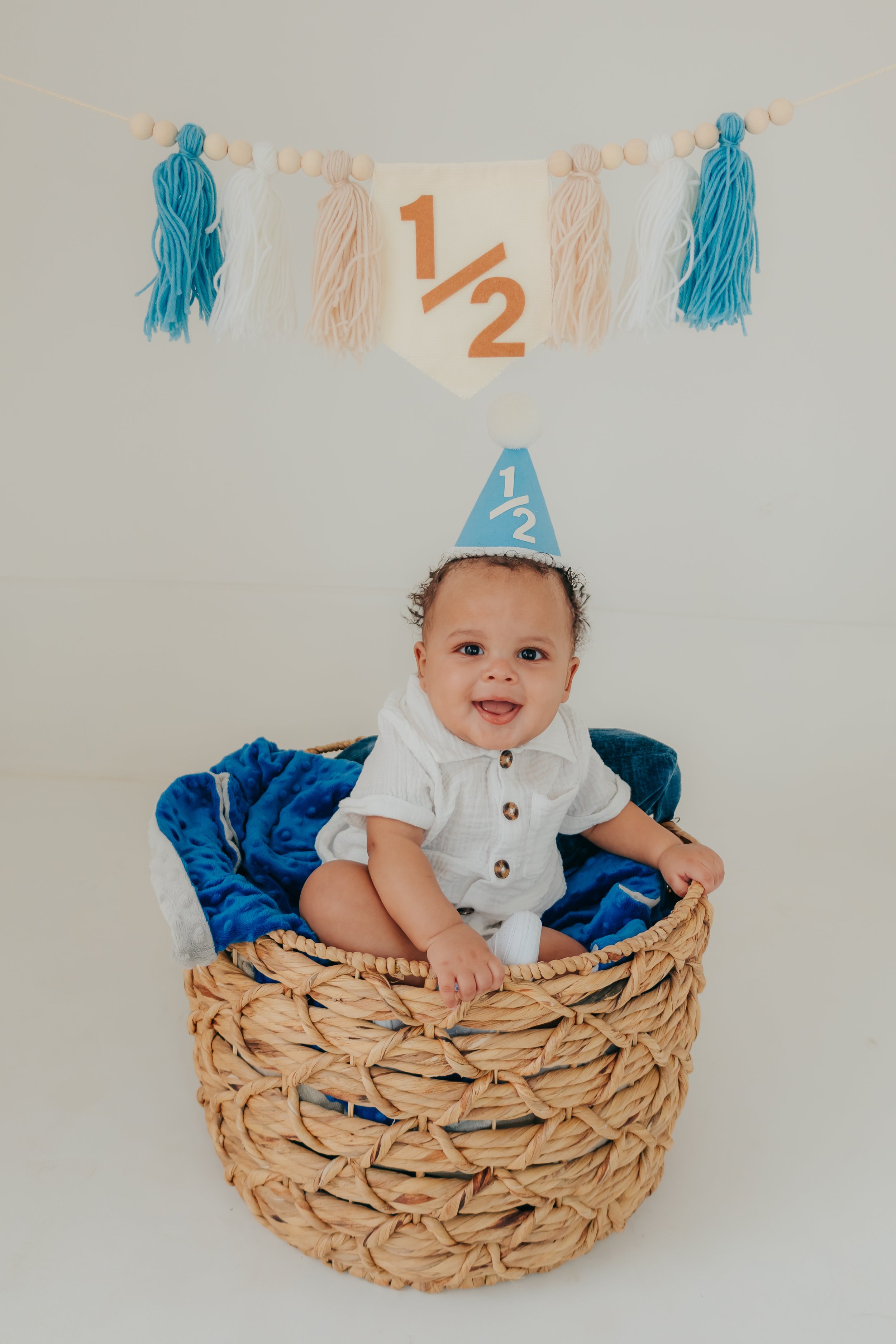 Smiling baby sitting in a basket, wearing a blue birthday hat with a white pom-pom, celebrating a half-year milestone with a "1/2" banner and tassel decorations in the background. Memphis and Hernando Family Photographer.