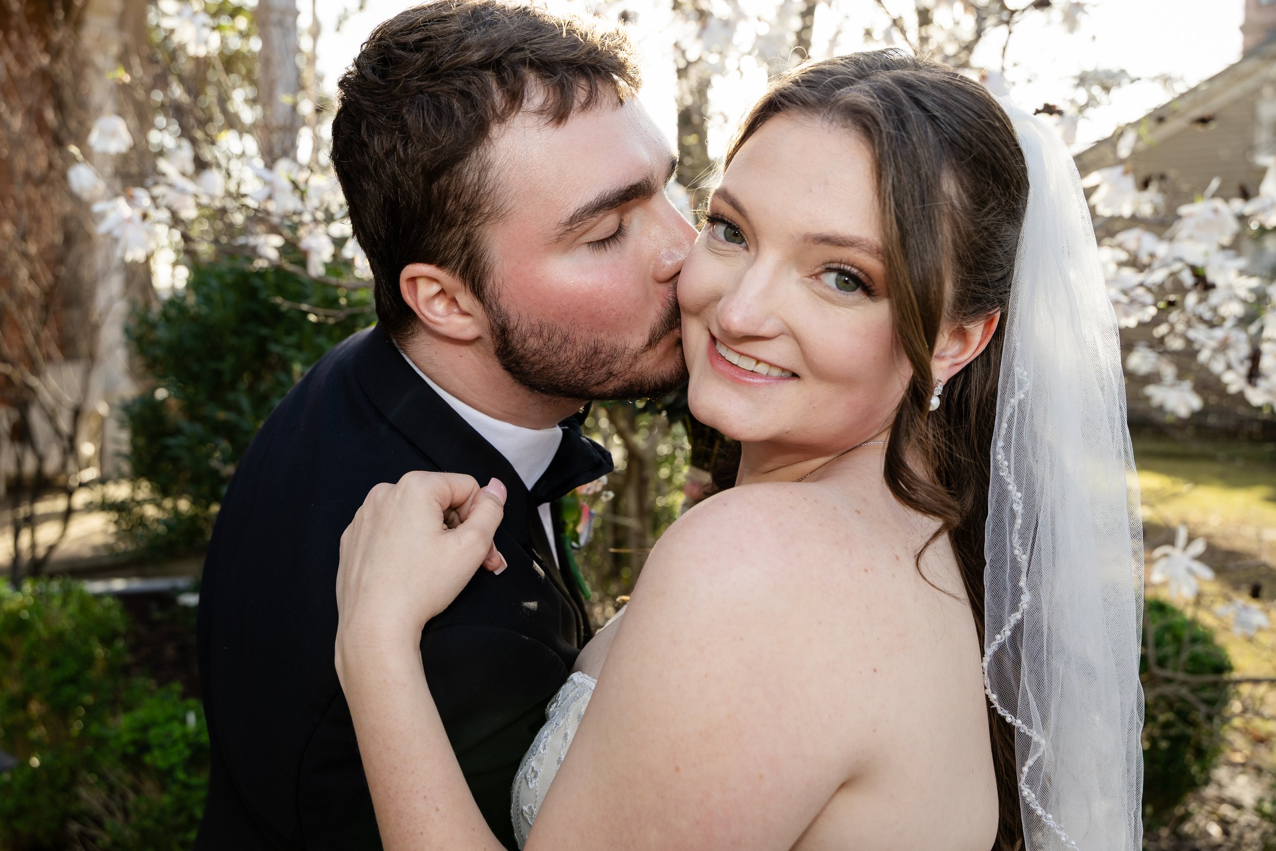 A happy bride and groom embrace outdoors; the groom is kissing the bride on her cheek, and she is smiling at the camera, with blooming trees in the background. Woodruff Fontaine museum wedding photographer