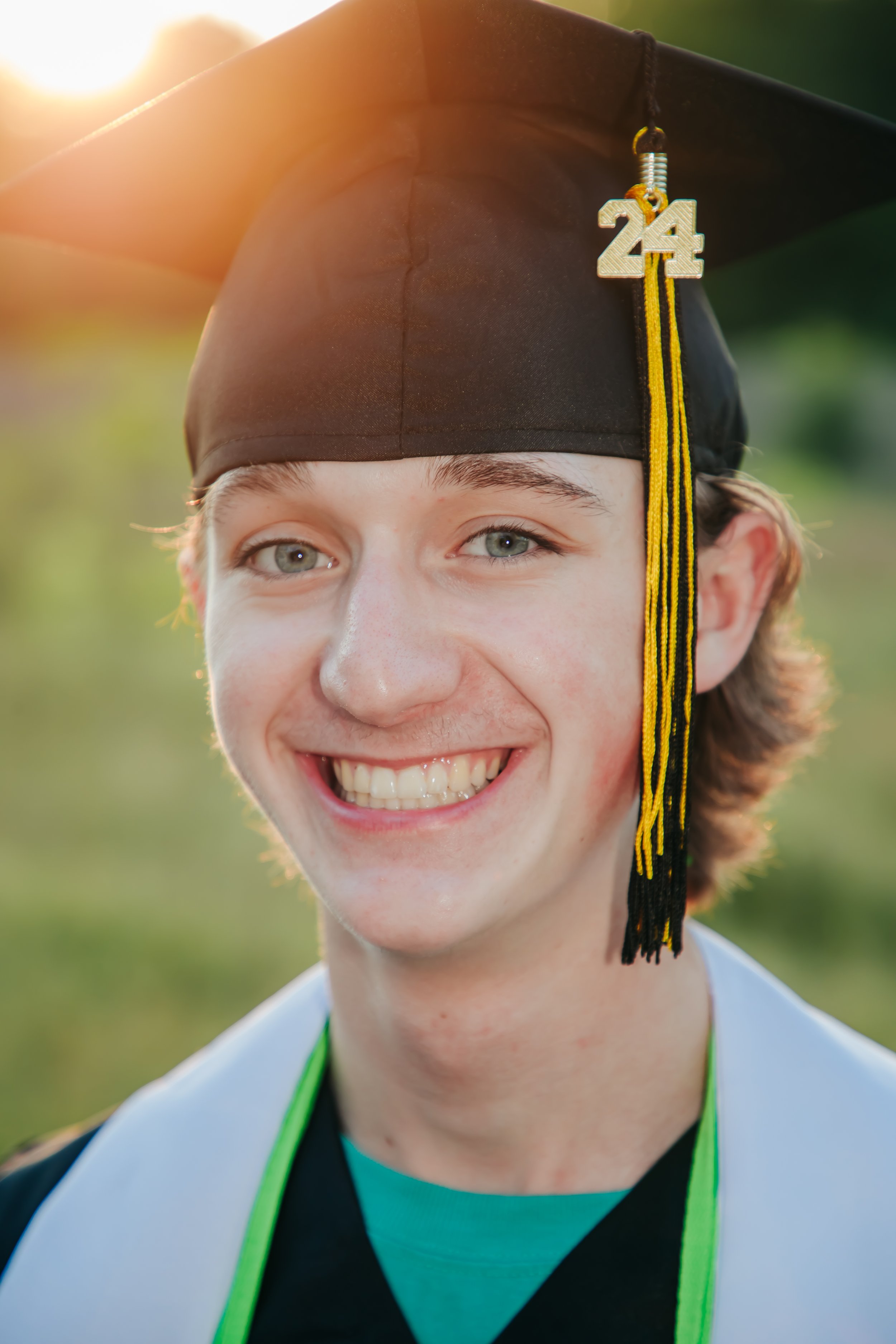 A young man in a graduation cap with a gold and black tassel and 24 charm, smiling outdoors in sunlight. Hernando, Memphis, Olive Branch, Nesbit senior photographer.