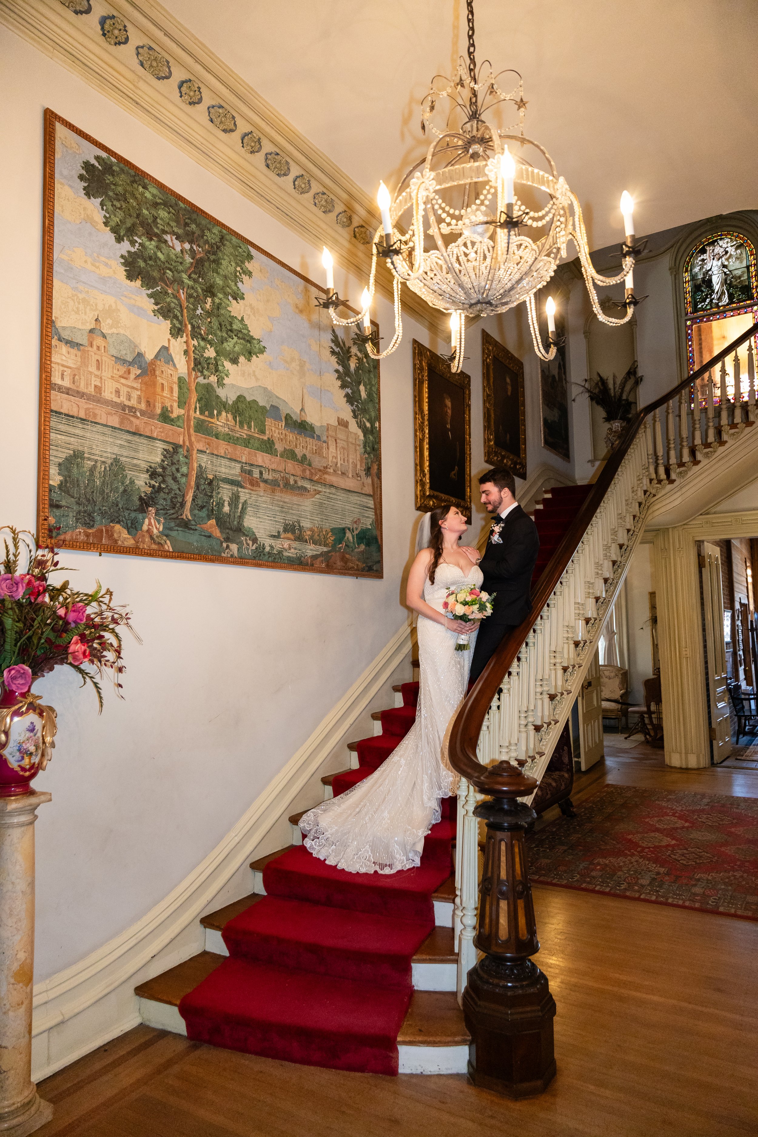 Bride and groom standing on a curved, red-carpeted staircase inside a grand, vintage mansion, looking at each other. Woodruff Fontaine Museum Wedding photographer.