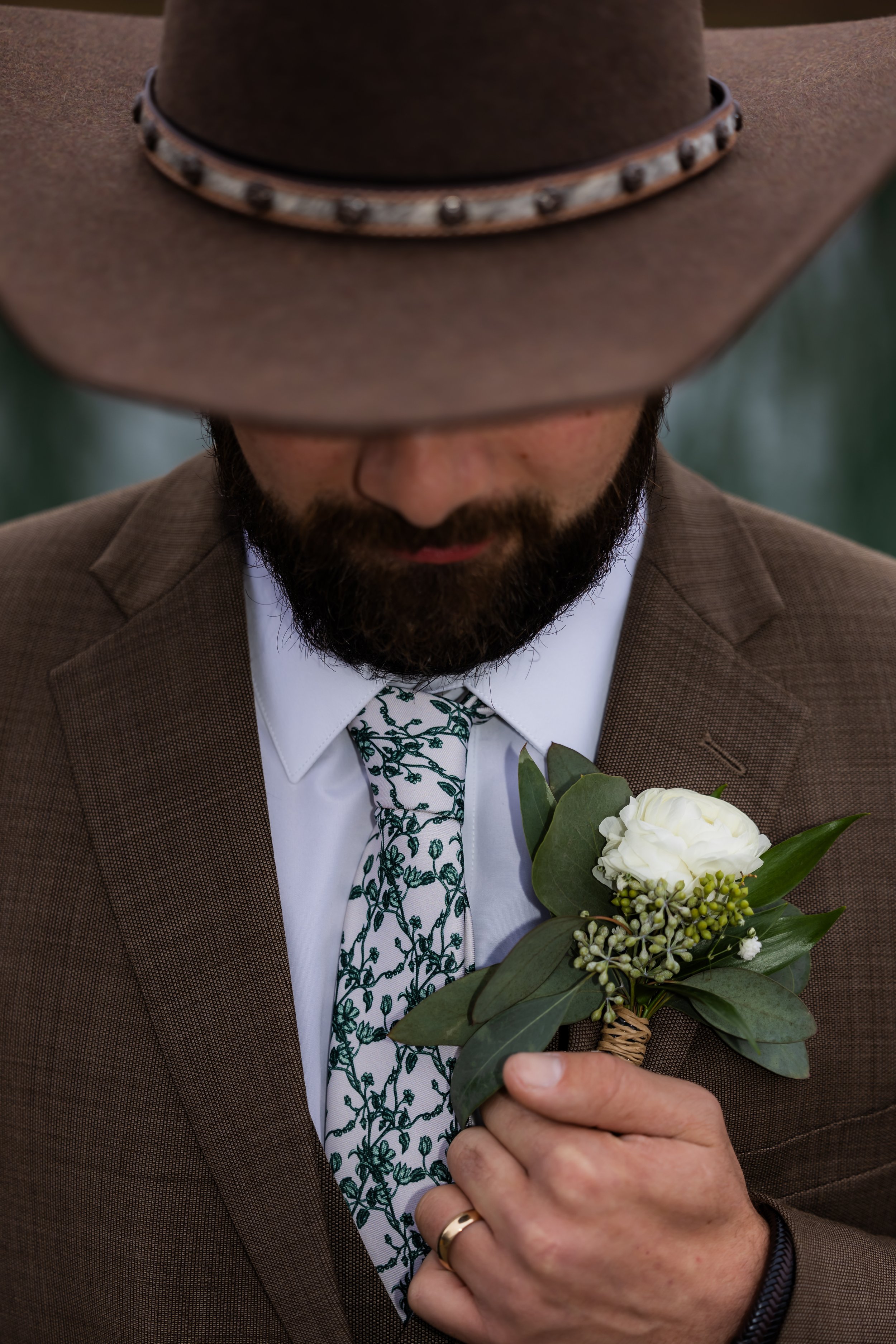 A man wearing a brown fedora hat, brown suit jacket, and a floral tie, holding a white boutonniere made of flowers and greenery against his chest. Robinshaw Wedding photographer Tennessee.