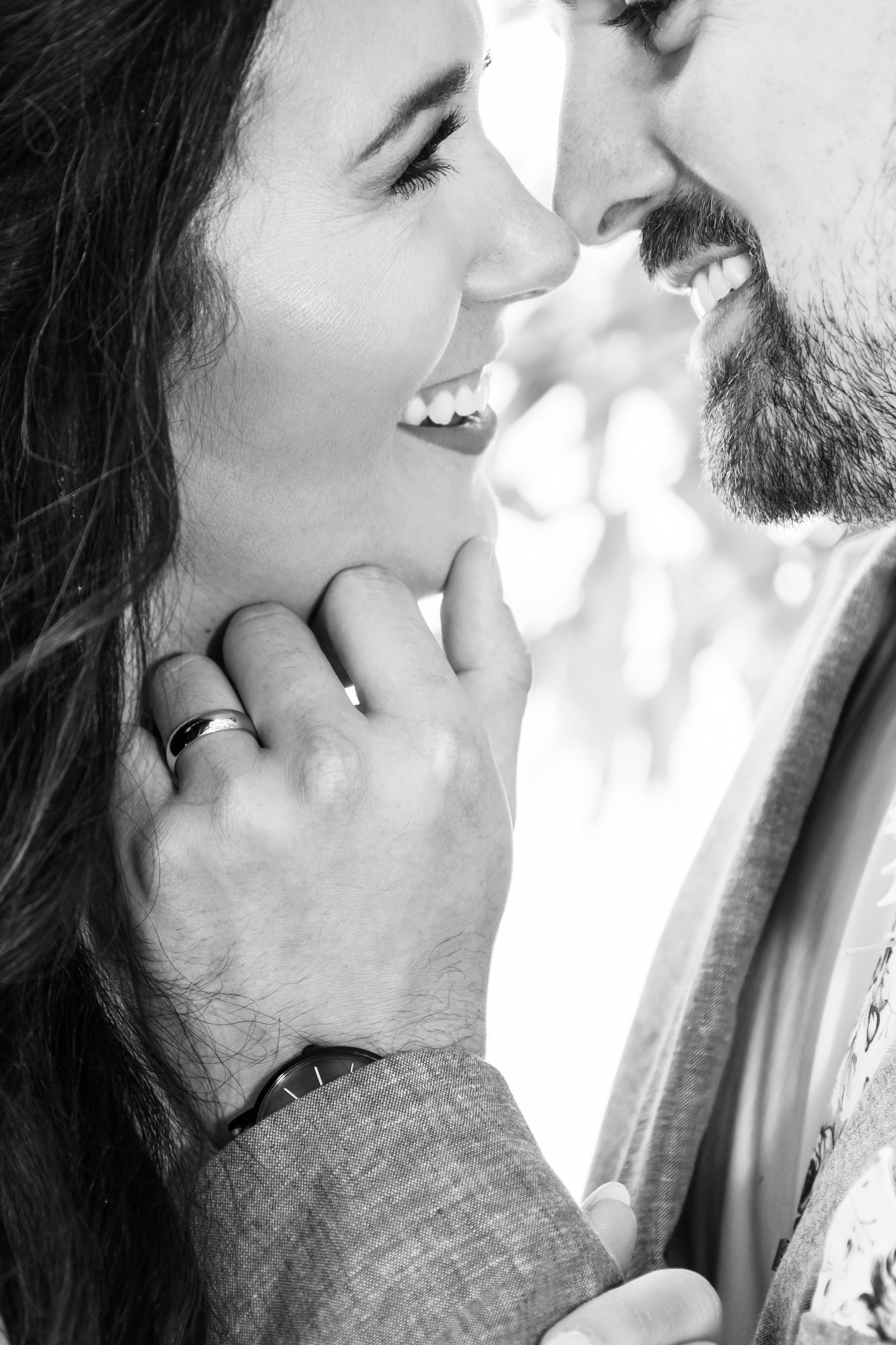 A close-up black and white photo of a man and woman smiling and touching foreheads. The woman has long dark hair and is wearing makeup, while the man has a beard and mustache, wearing a suit jacket and a watch. Hedgefarms wedding photographer.