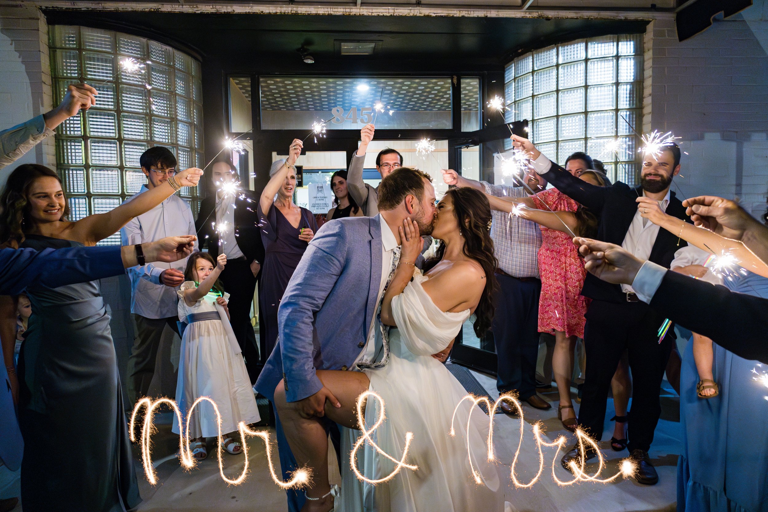 A newlywed couple sharing a kiss at their wedding reception while friends and family hold sparklers around them, with "MOL & MUE" written in sparklers at the bottom. Memphis wedding photographer. Soul and Spirit Brewery