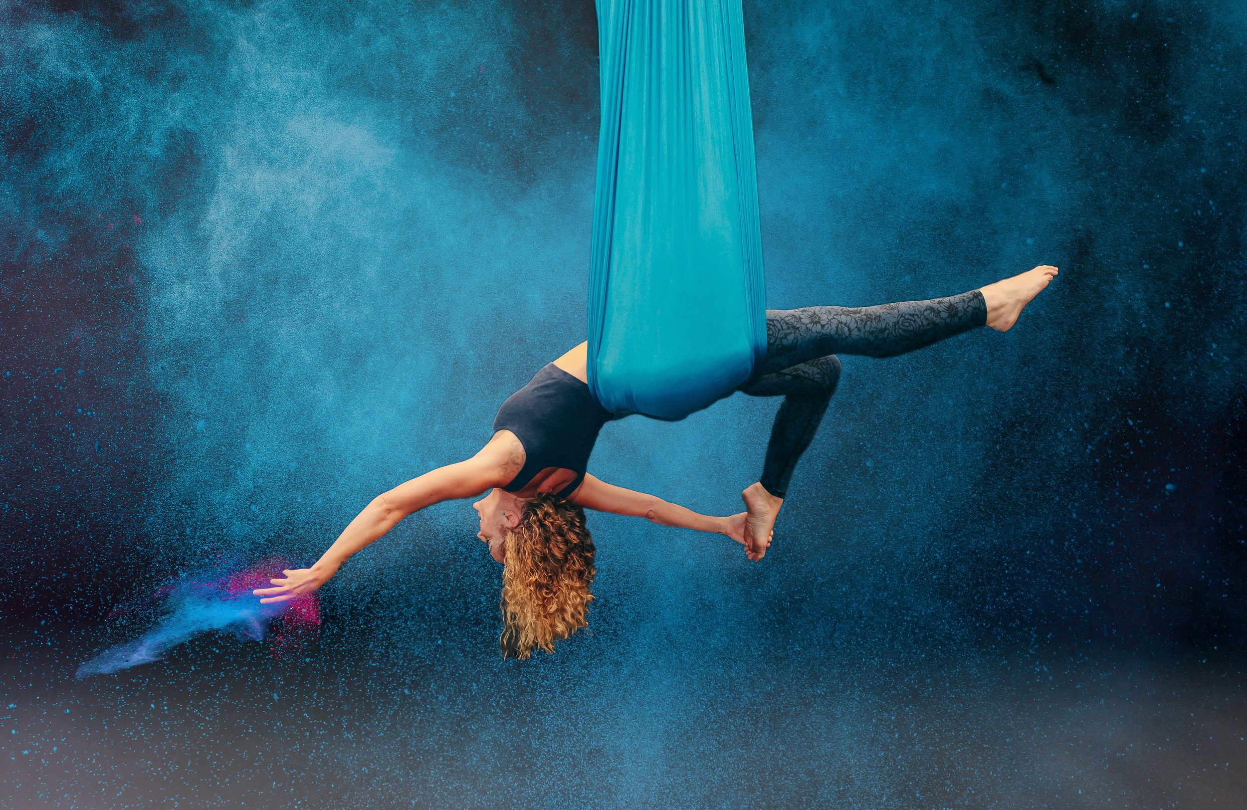 A woman performing an aerial silk acrobatics move suspended upside down in a blue silk fabric amid colorful powder clouds. Business Branding photographer Hernando Mississippi.