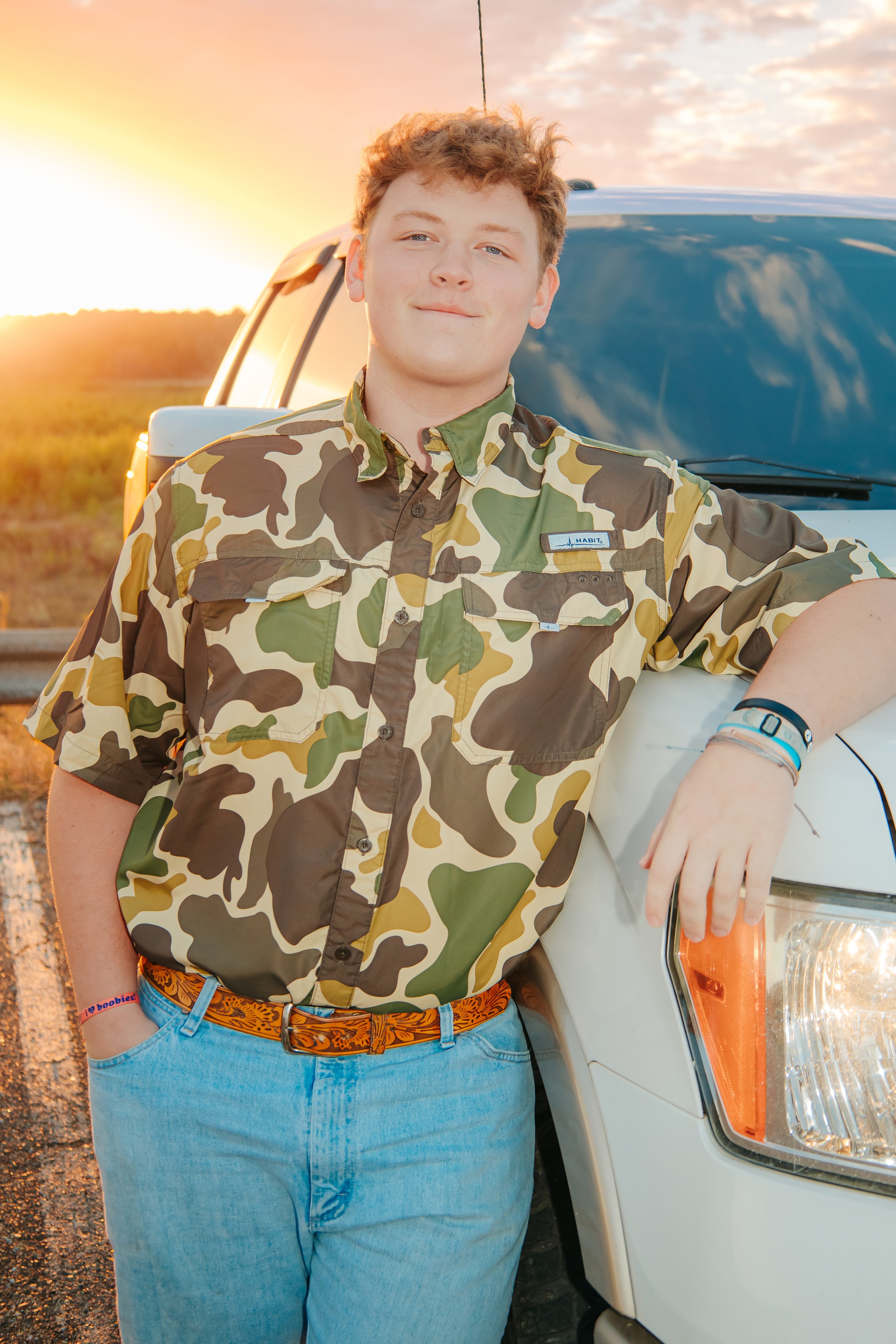 Young man leaning on a white vehicle with a sunset in the background, wearing a camouflage shirt and jeans. Senior Photography in Hernando, Memphis, and Olive Branch.