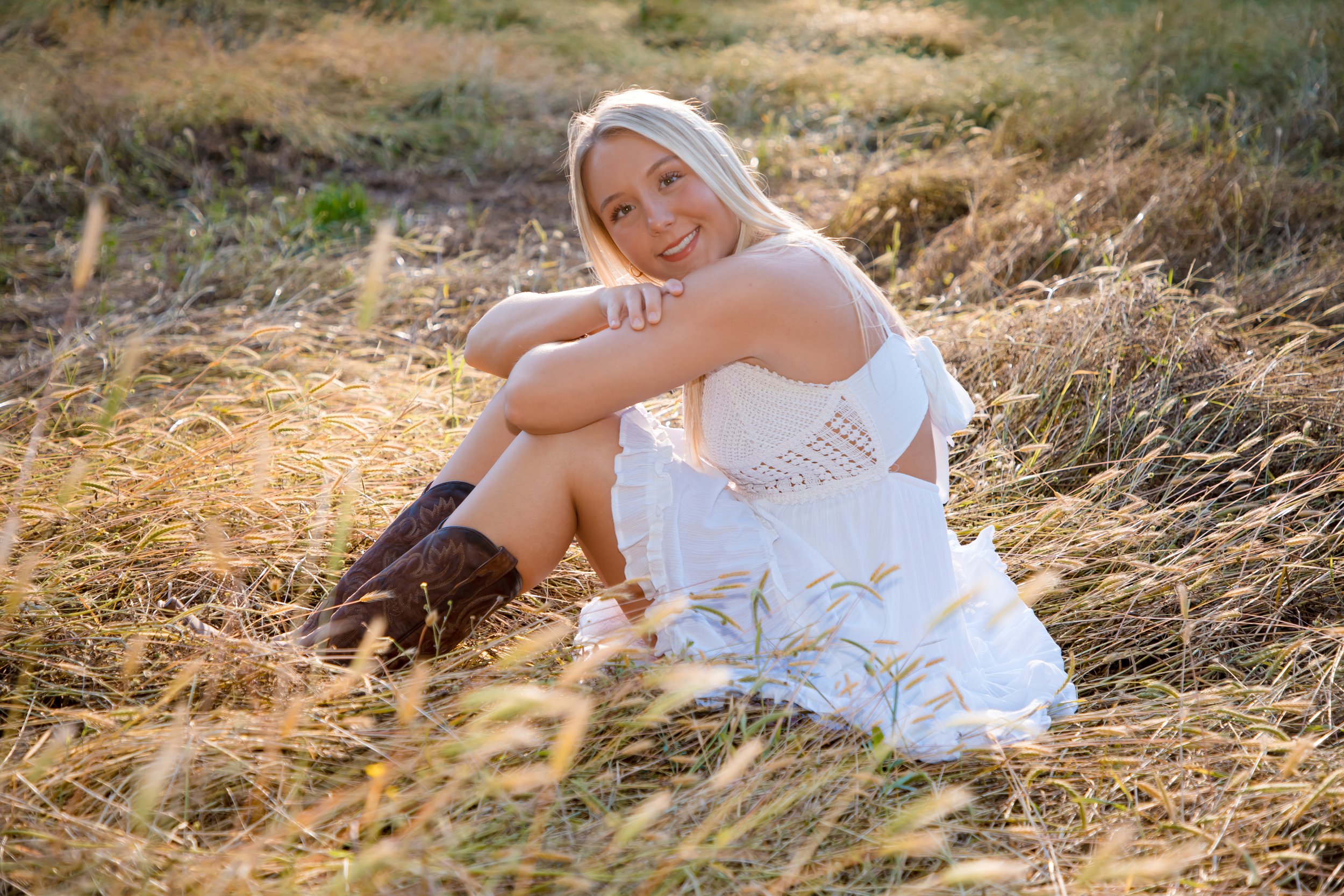 A young woman in a white dress sitting in a grassy field with golden sunlight, smiling and looking at the camera. Senior Photography in Hernando.
