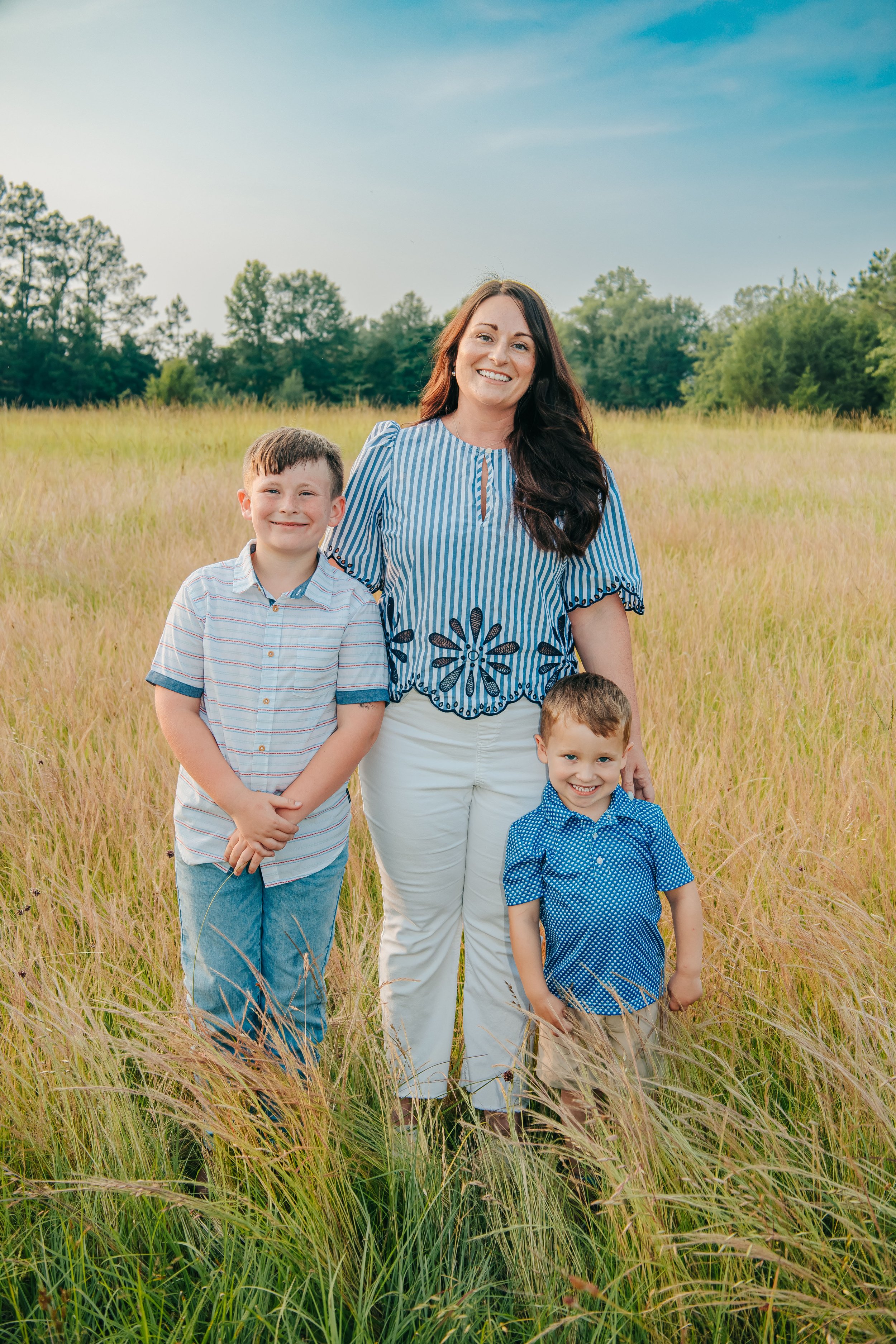 A woman with two young boys standing in a grassy field with trees in the background, smiling at the camera. Hernando, Memphis, and Olive Branch family photographer