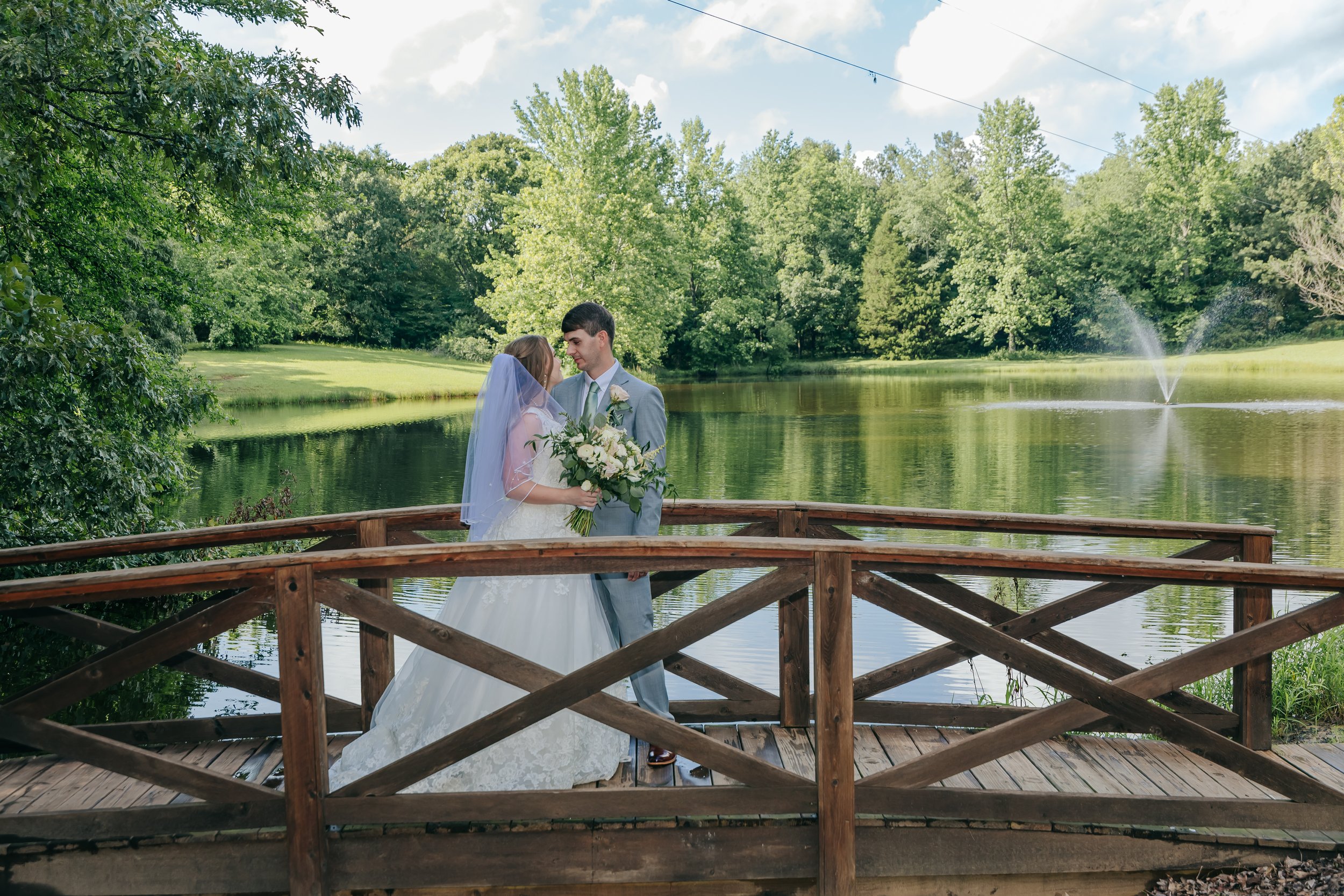 A bride and groom standing on a wooden bridge by a lake, surrounded by lush green trees, during their wedding. The bride holds a bouquet, and they are looking at each other affectionately. Kincaid Manor Wedding photography. Holly springs Mississippi.