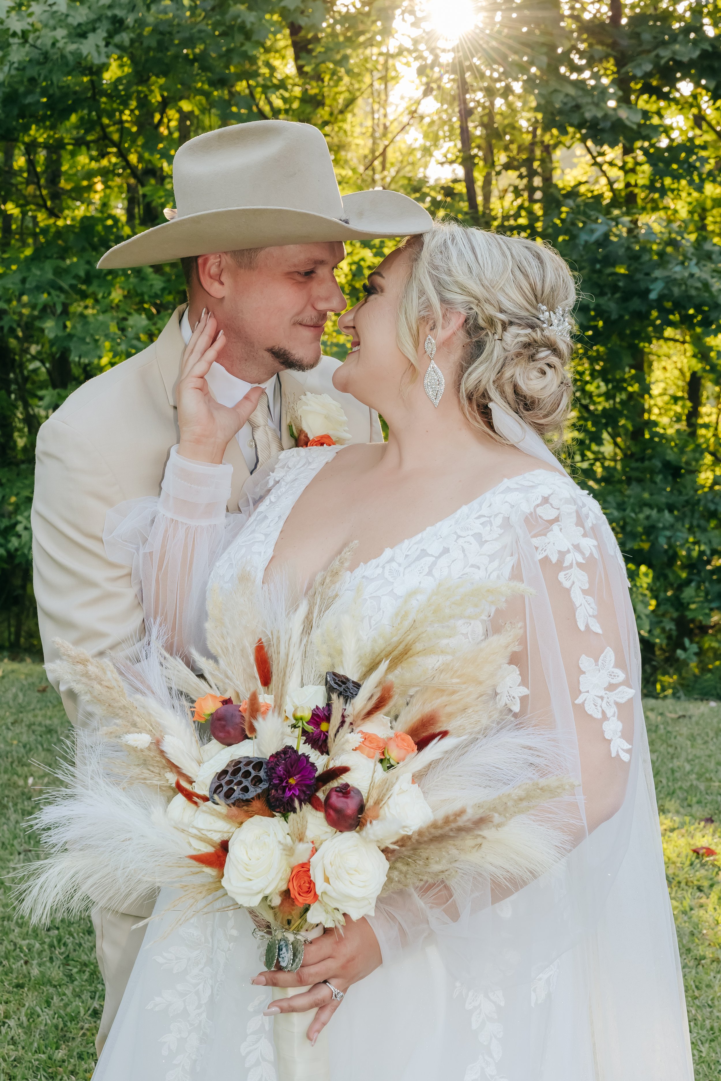 Bride and groom sharing a close moment outdoors during their wedding, with the groom wearing a beige cowboy hat and the bride holding a bouquet of flowers and dried plants. Tennessee Wedding Photographer. Yellowheart barn.