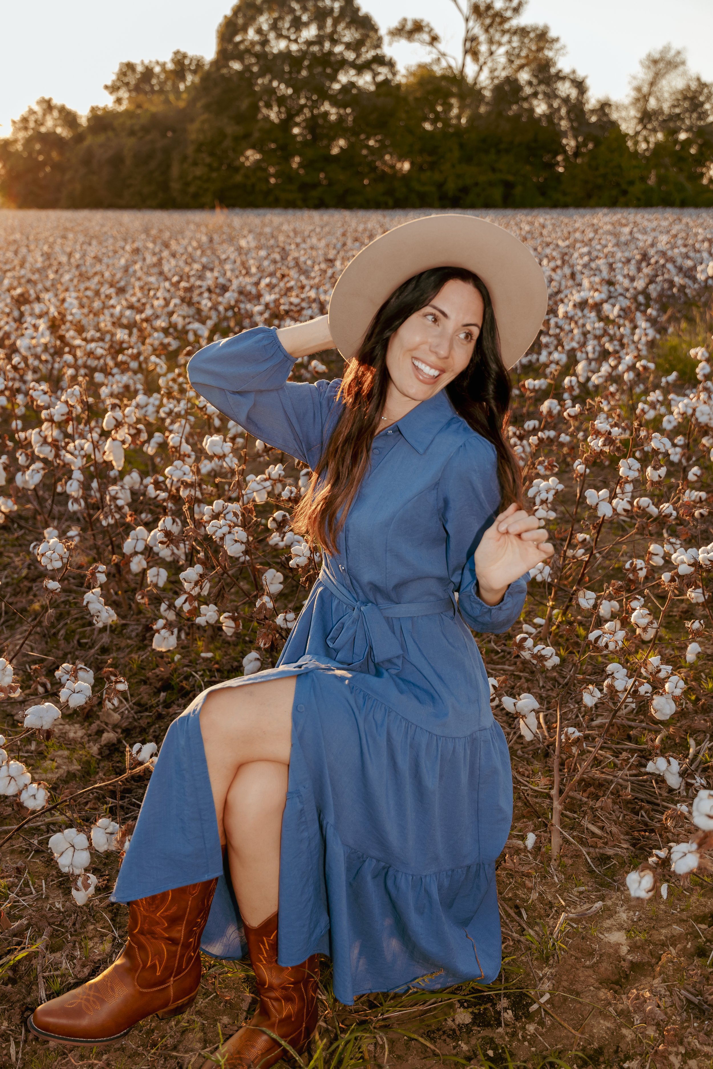 A woman in a blue dress and cowboy boots, wearing a wide-brimmed hat, is smiling and posing in a cotton field during sunset. Family portrait photographer Hernando Mississippi.