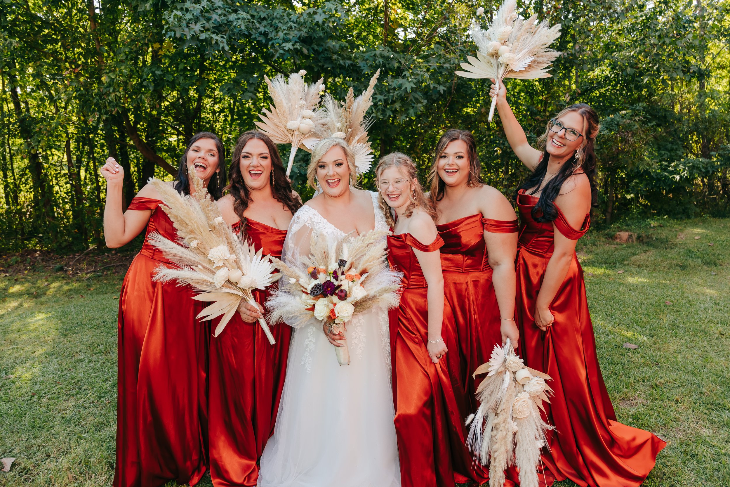 A bride and six bridesmaids smiling and celebrating outdoors in a park with green trees. The bride is wearing a white dress, holding a bouquet, and the bridesmaids are wearing matching red dresses, holding dried floral bouquets and fans. Yellowheart