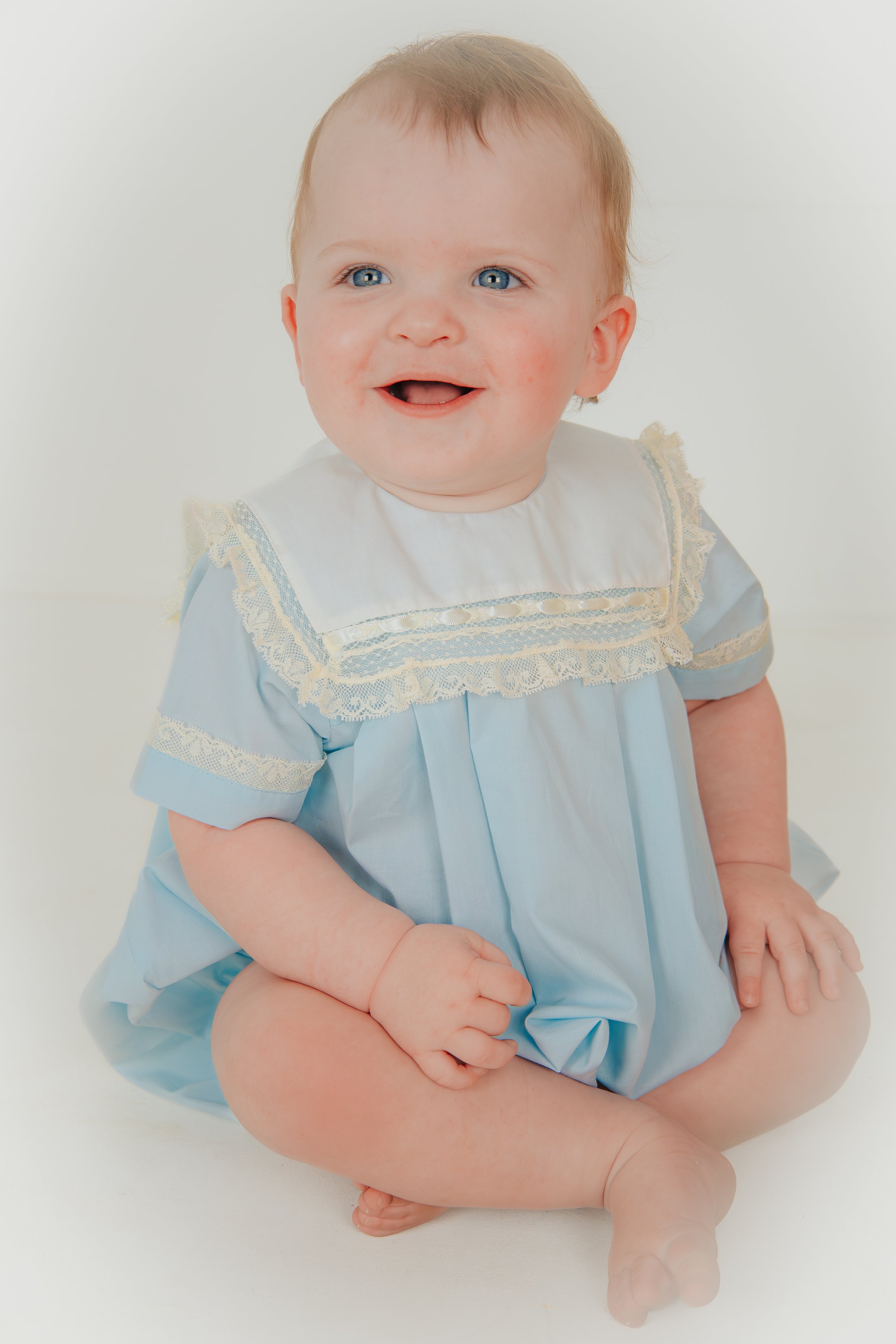 Smiling toddler with blue eyes, light brown hair, wearing a light blue dress with lace details, sitting on a white surface. Heirloom Child Photography. Memphis and Hernando Family Photographer.