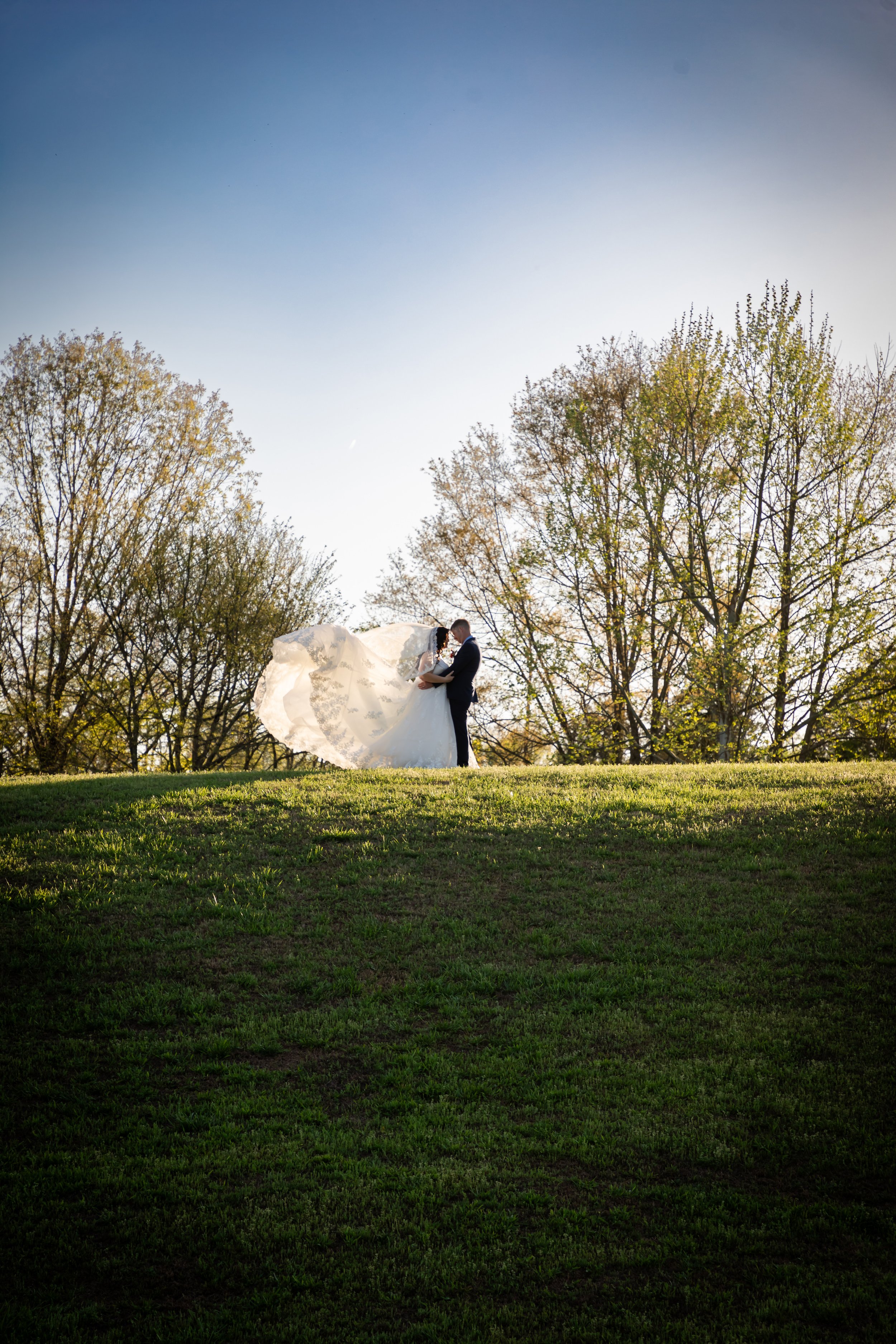A bride and groom standing on a hill at sunset, with trees in the background and the bride's veil flowing in the wind. Wedding Photographer in Tennessee. Robins Nest.