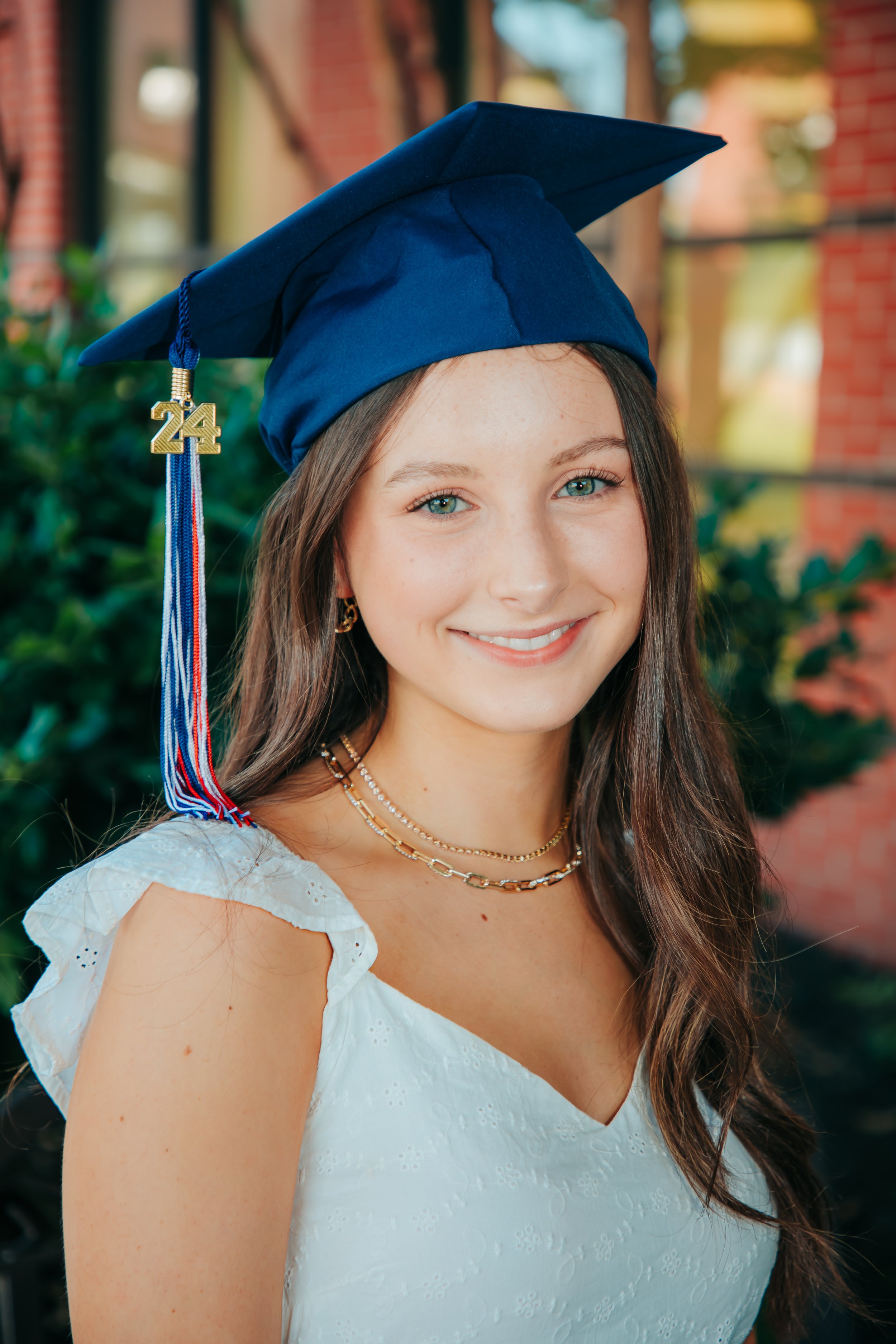 A young woman with long brown hair and blue eyes wearing a blue graduation cap with a gold '24' charm, a white eyelet dress, and layered gold jewelry, smiling outside in front of green bushes and a brick building. Hernando, Memphis, and Olive Branch.