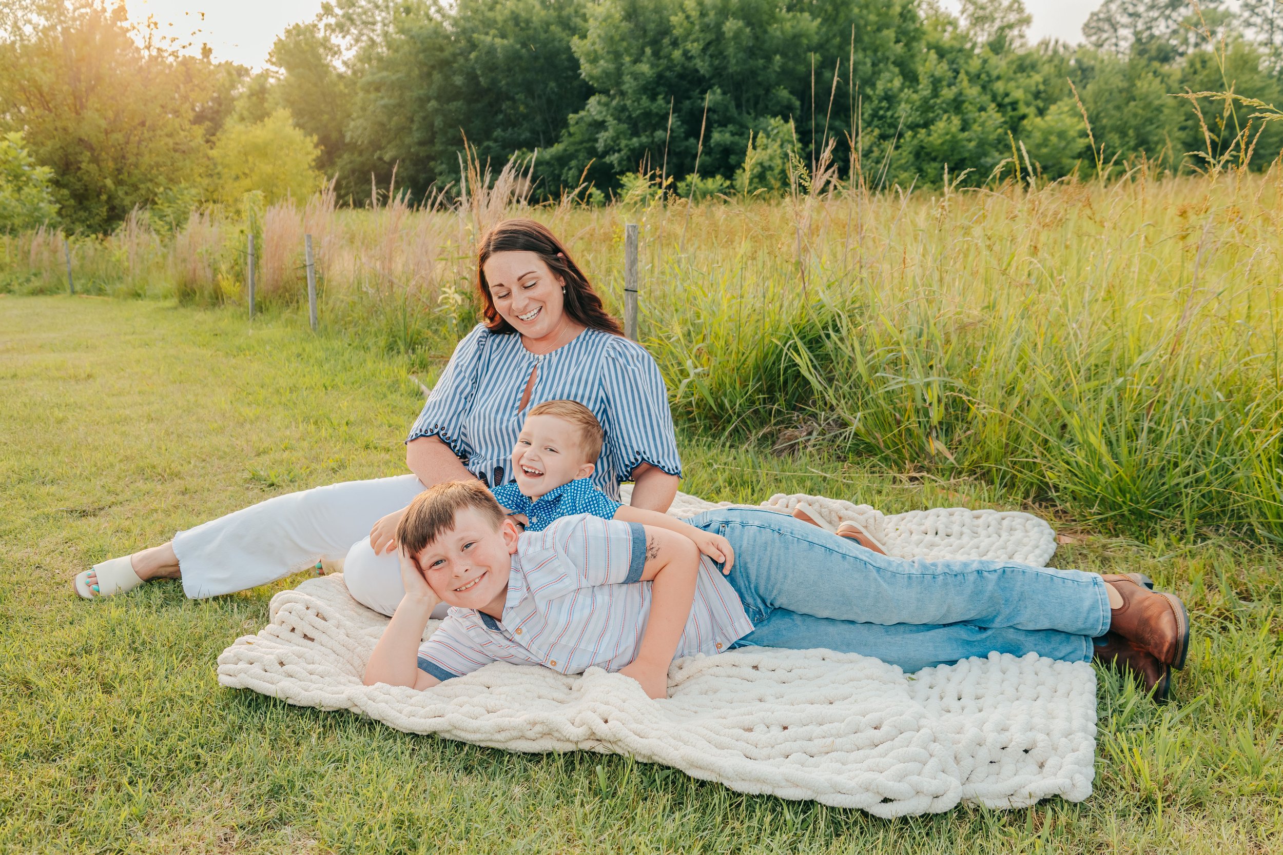 A woman and two young boys relaxing on a blanket outdoors in a grassy field with tall grass and trees in the background, smiling and enjoying a sunny day. Hernando, Memphis, and Olive Branch family photographer.