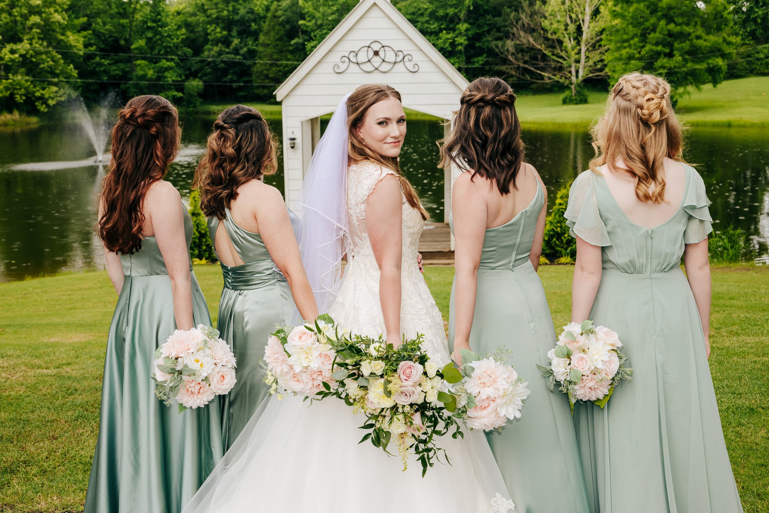 Bride in white wedding dress with veil and five bridesmaids in matching sage green dresses holding bouquets, standing outdoors near a pond with a small white chapel in the background. Kincaid Manor. Hernando Wedding Photographer. Holly Springs