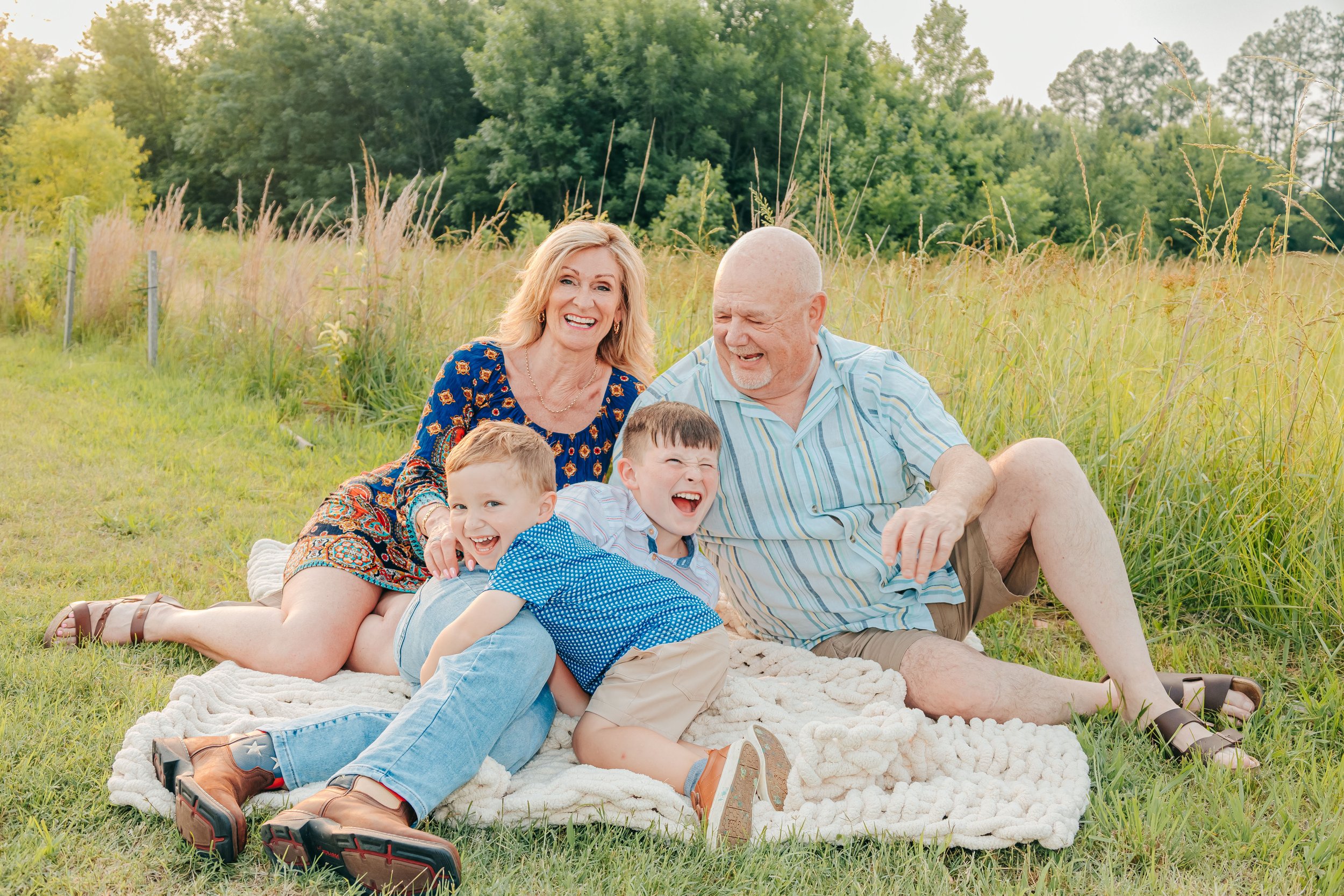 A happy family of five, including two children and two older adults, lying on a blanket outdoors in a grassy field with trees in the background, smiling and laughing together.