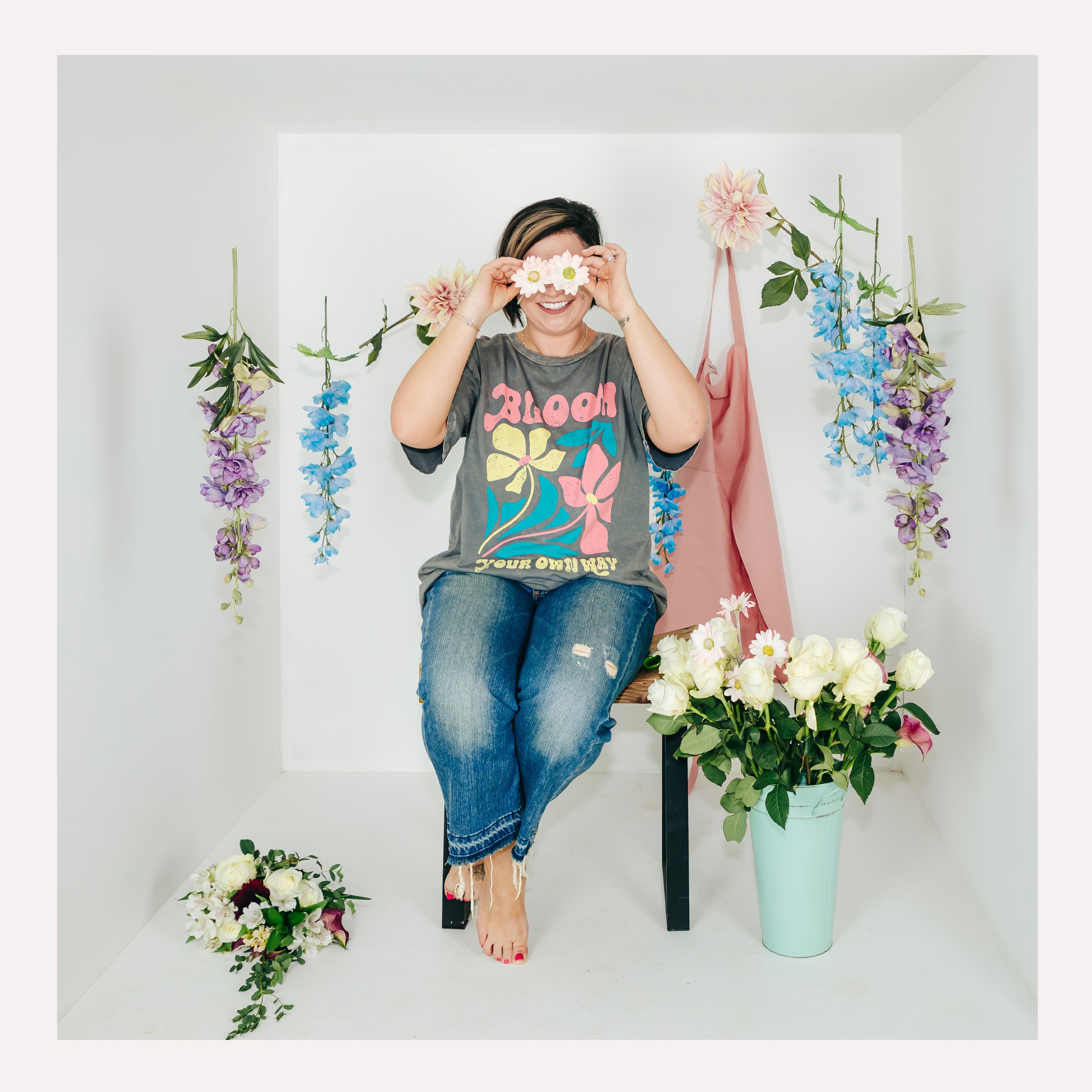 Woman sitting on a black stool in a small white room decorated with hanging purple and blue flowers, holding a pair of white flower glasses in front of her eyes, smiling, with large white roses in a light blue vase beside her and a small bouquet of f