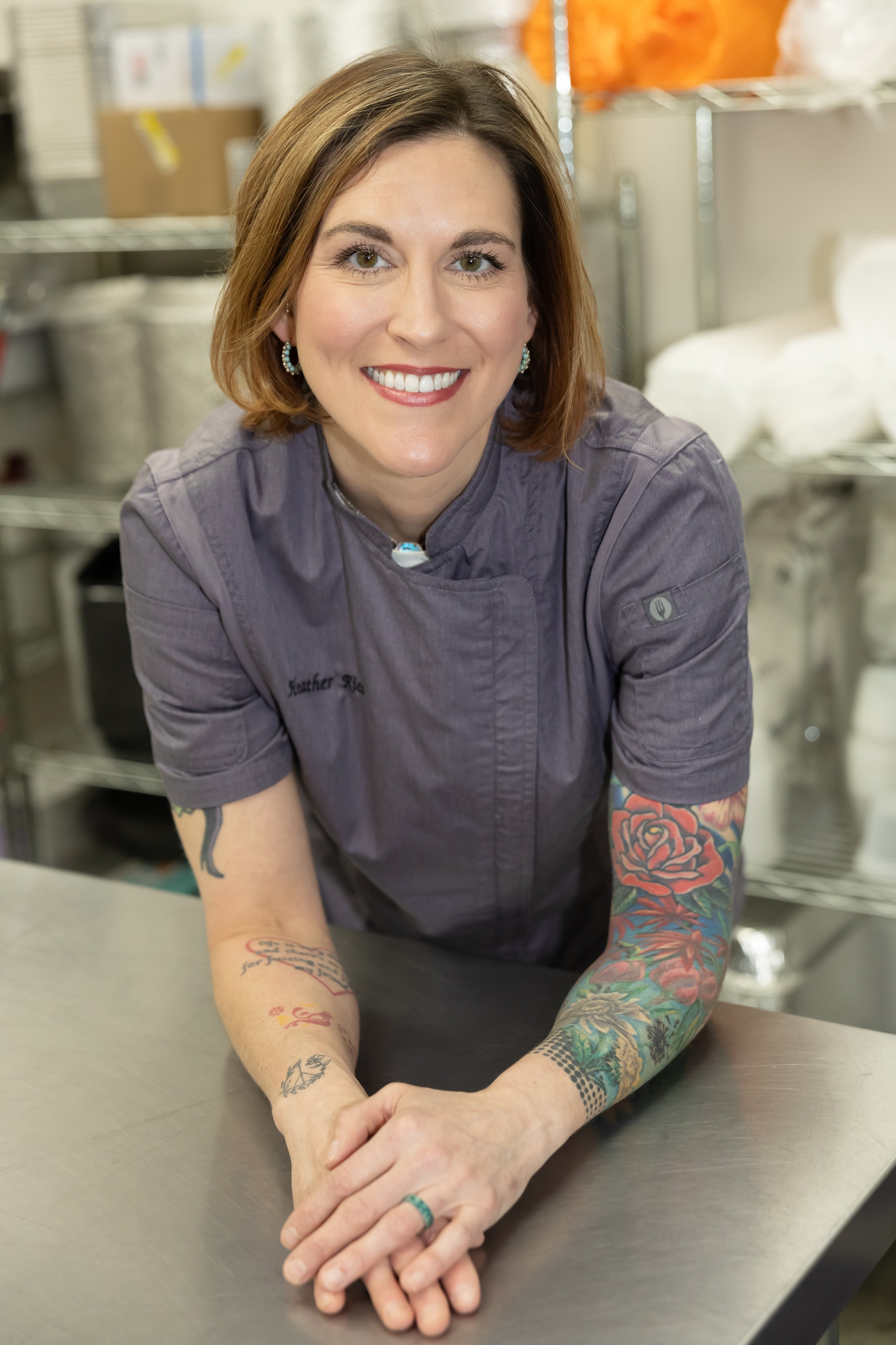 A woman with short brown hair and tattoos wearing a gray chef's jacket, smiling and leaning on a stainless steel counter in a commercial kitchen, with shelves and kitchen supplies in the background. Memphis, Hernando, business branding photographer.