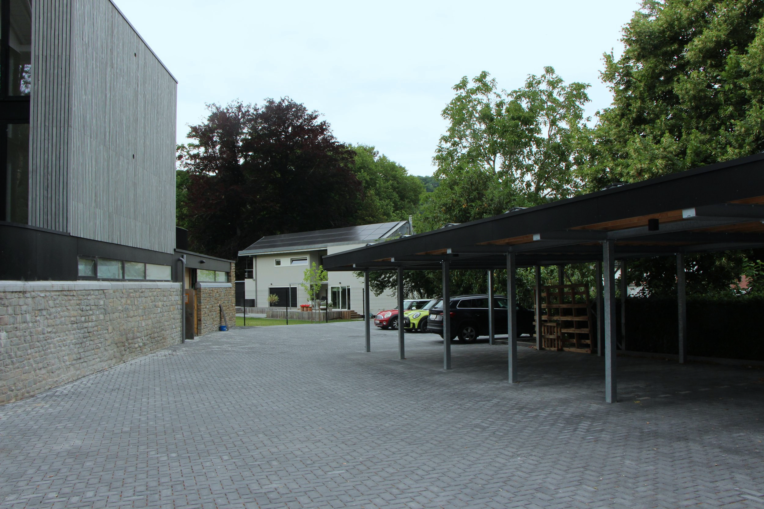 Un parking extérieur vide avec quelques voitures sous un parking en structure métallique, des arbres verts en arrière-plan, côté bâtiment moderne en pierre et bois.