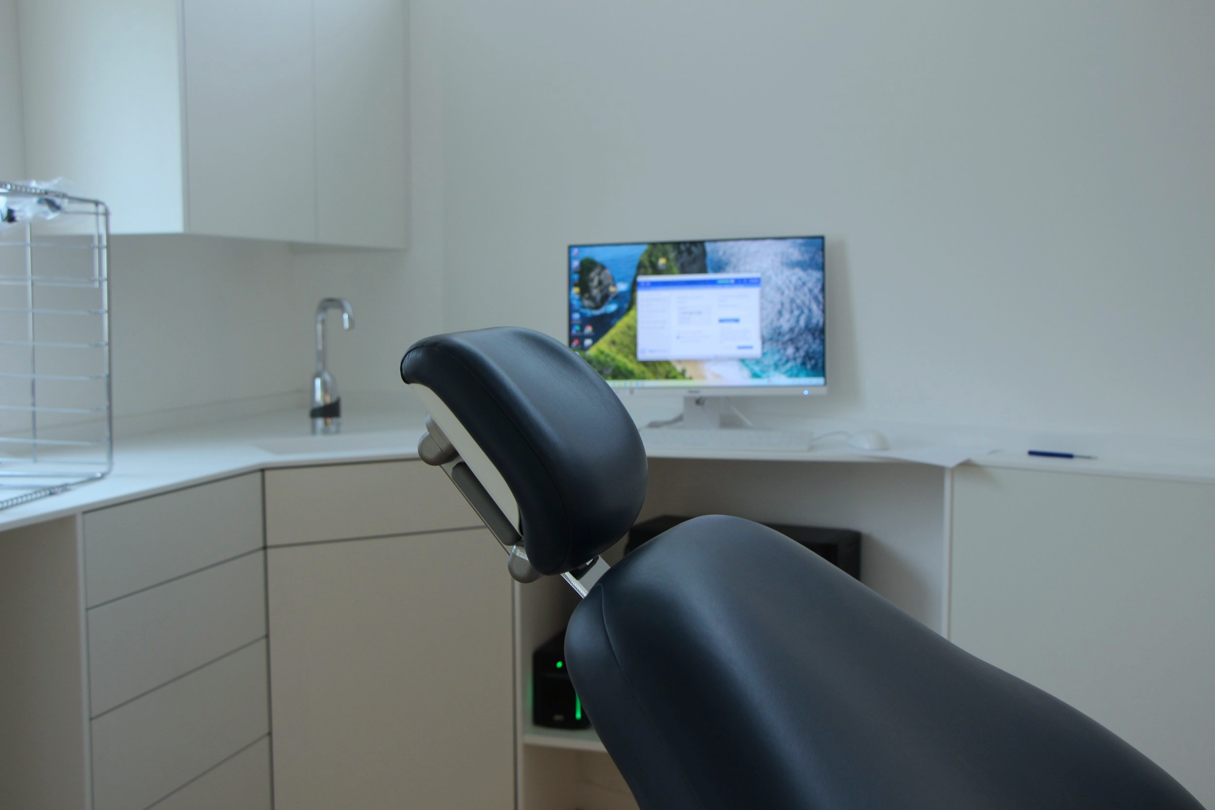 Salle de consultation médicale avec un fauteuil dentaire ou médical, un bureau blanc, un ordinateur à écran large affichant un fond de paysage et une fenêtre de dialogue, un évier avec un robinet, des armoires blanches et un panier en métal.