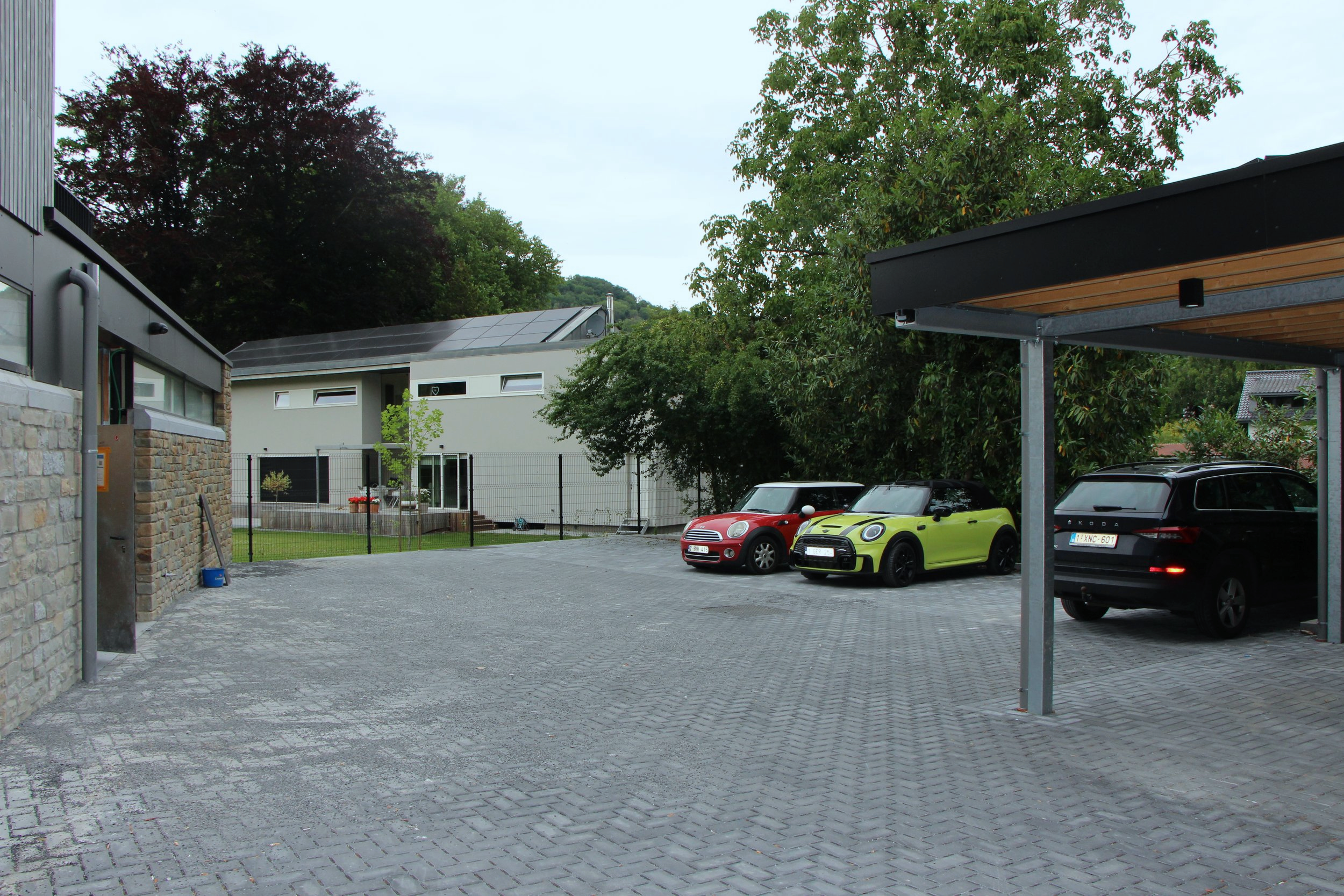 Un parking résidentiel en pierre grey avec trois voitures, dont une Mini Cooper rouge, une Mini Cooper vert lime, et une stationnement couvert en bois, entouré de maisons modernes et d'arbres.